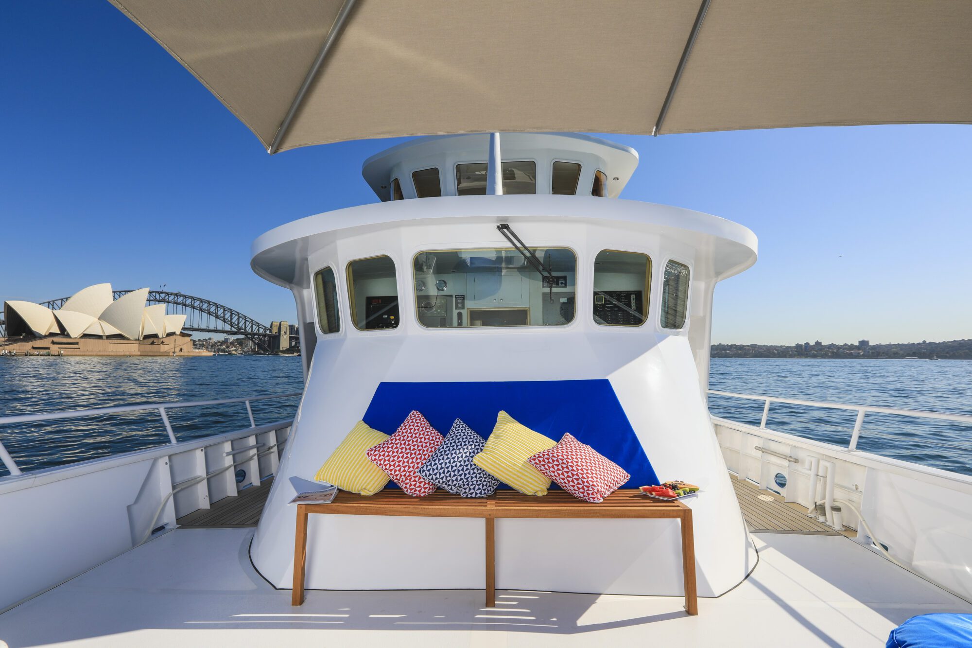 A yacht deck with colorful pillows on a bench under an umbrella, overlooking Sydney Opera House and Harbour Bridge on a sunny day.