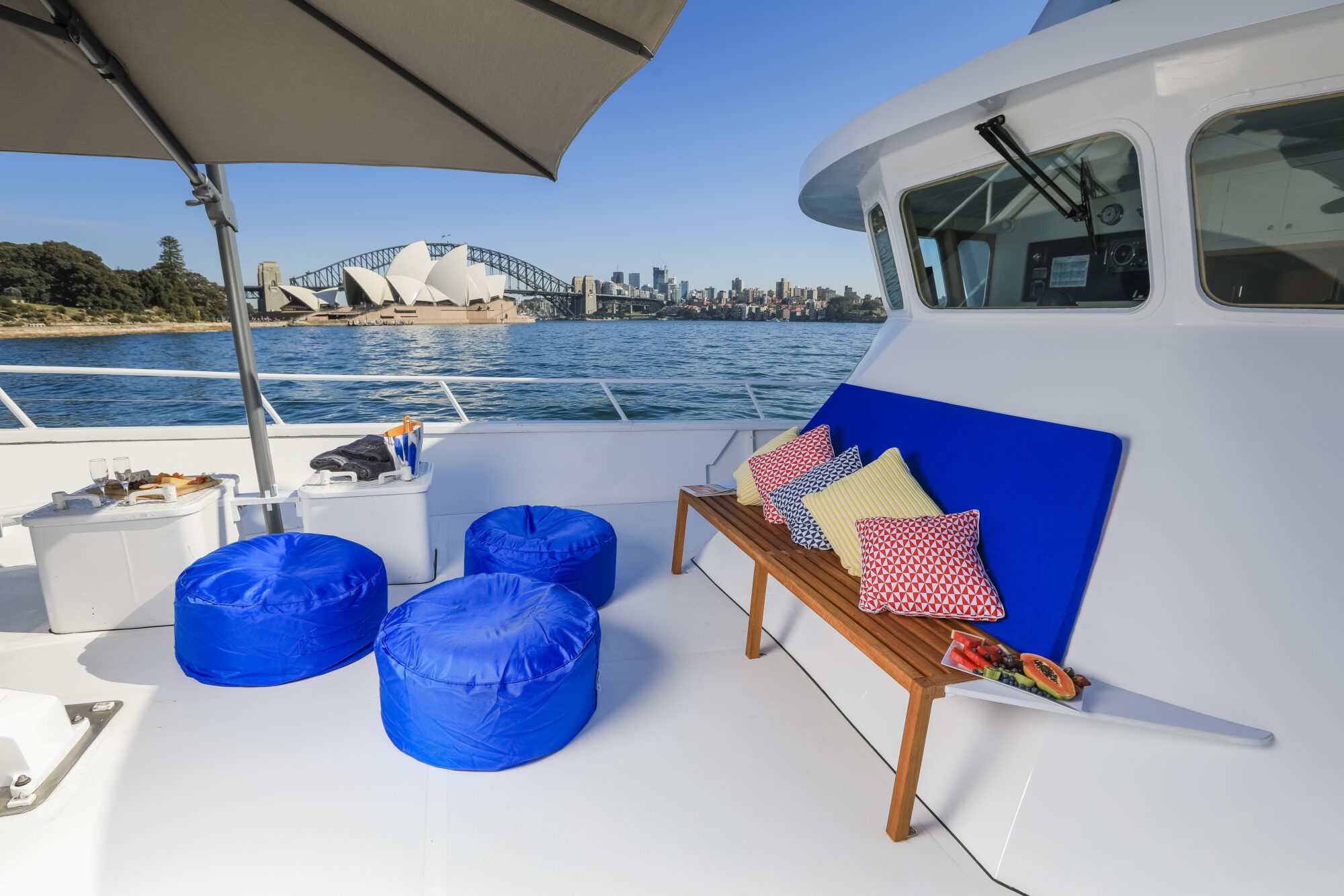 A yacht deck with blue beanbag chairs, a bench with colorful pillows, snacks on a tray, and a view of the Sydney Opera House and Harbour Bridge across the water under a clear blue sky.