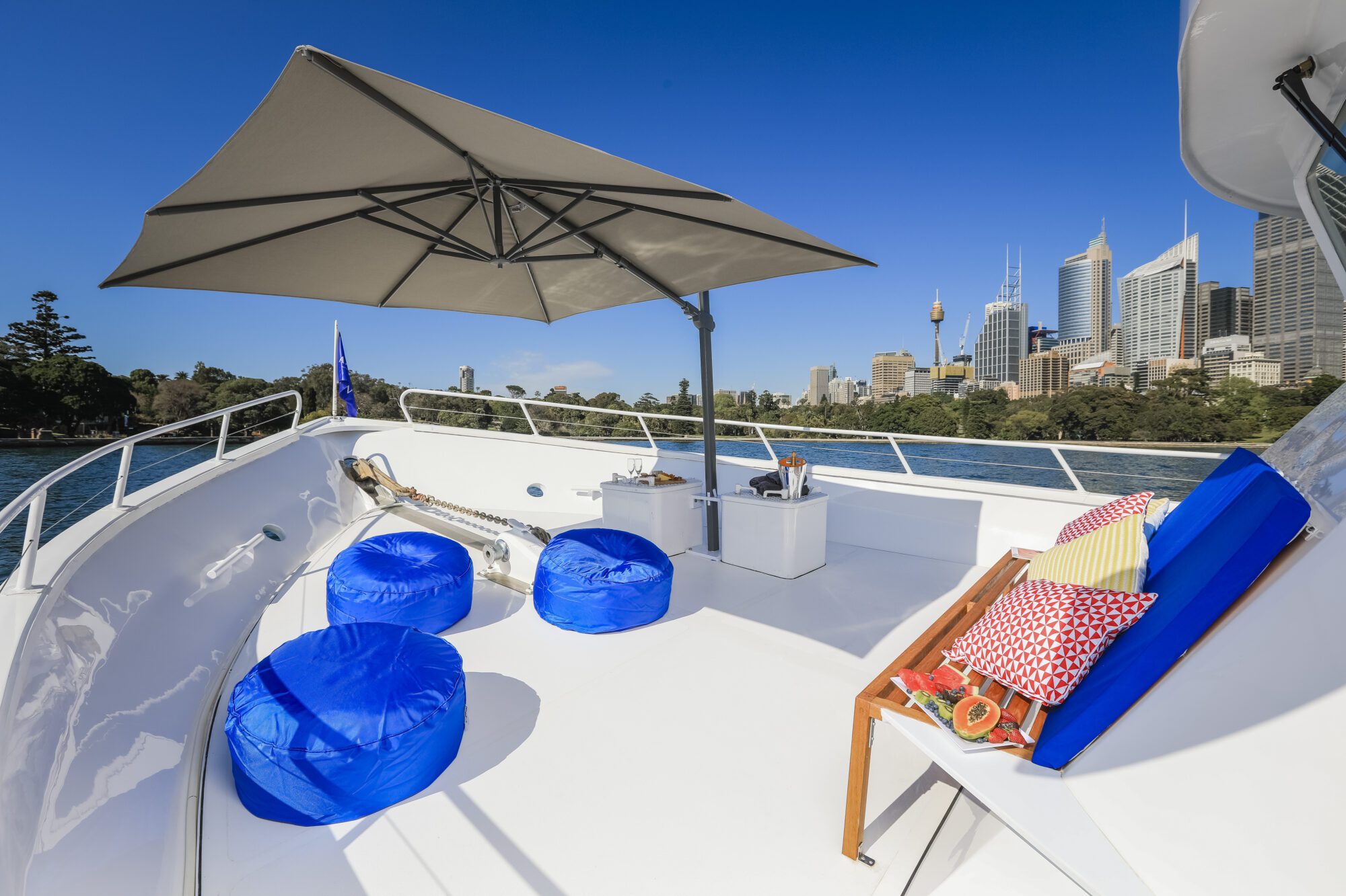 A sunny yacht deck with blue bean bag chairs, a cushioned bench with colorful pillows, and a large umbrella. The city skyline with tall buildings is visible in the background across the water.