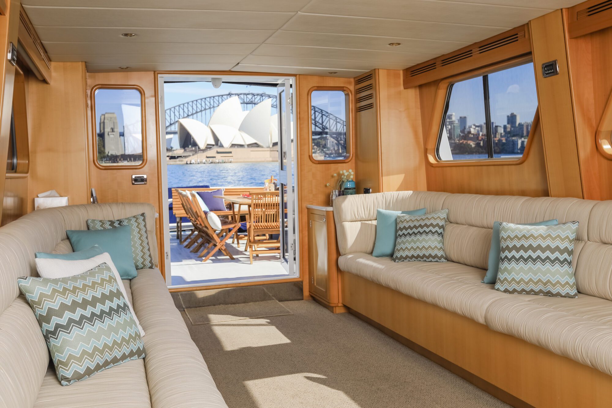 Interior of a yacht with beige sofas and patterned pillows, looking out through open doors to a deck with a dining table. The Sydney Opera House and Harbour Bridge are visible across the water outside.