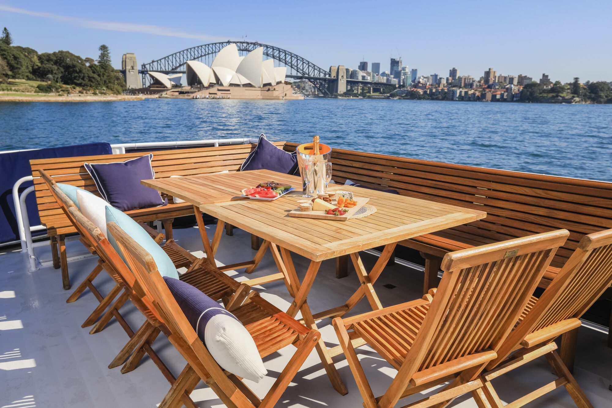 A wooden outdoor dining set with cushions sits on a boat deck overlooking Sydney Harbour, with the Sydney Opera House and Harbour Bridge visible in the background on a sunny day.