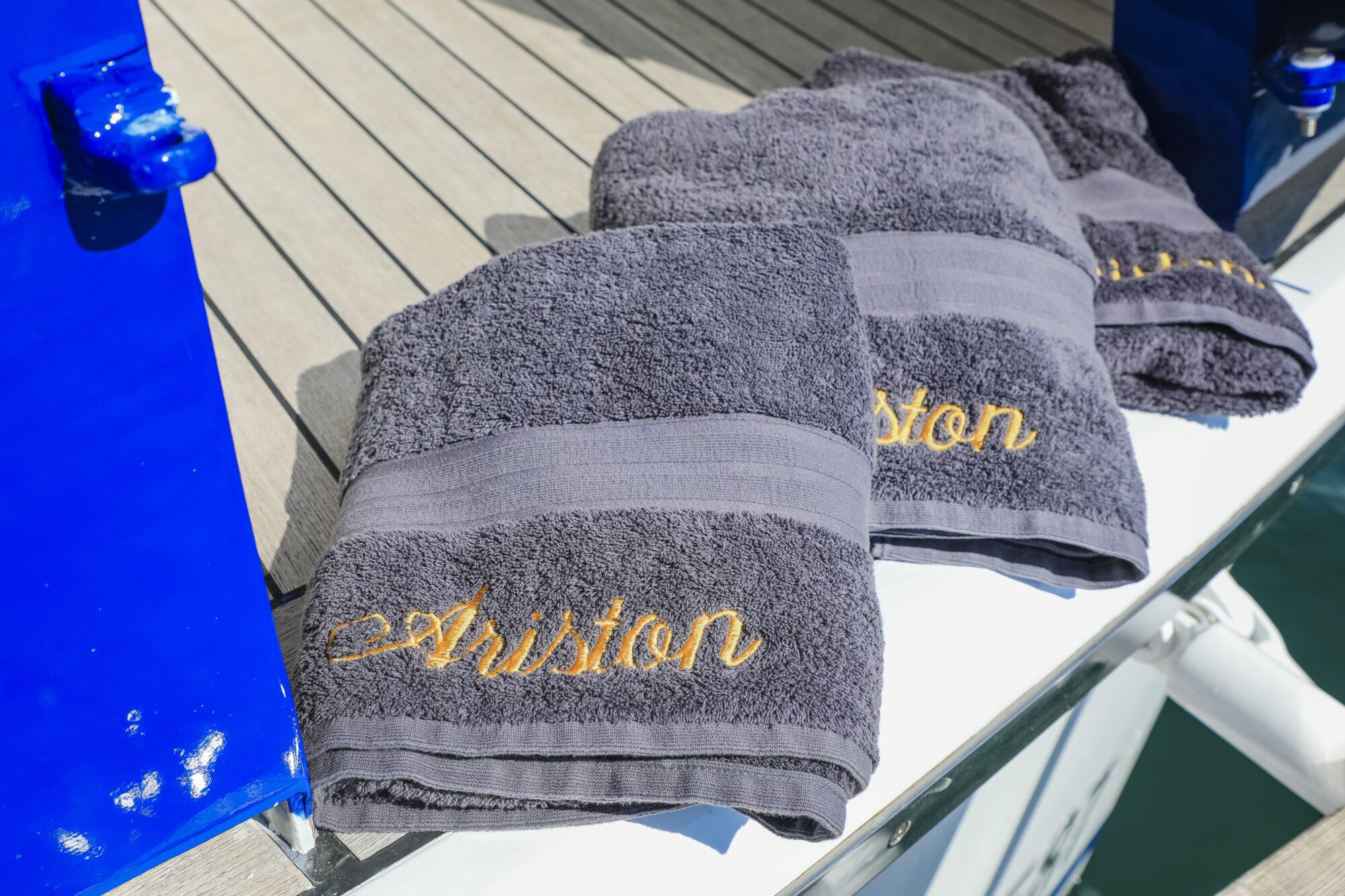 Three dark gray towels embroidered with the word “Ariston” in gold are neatly laid out on a wooden deck beside a blue railing, with sunlight casting shadows across the scene.