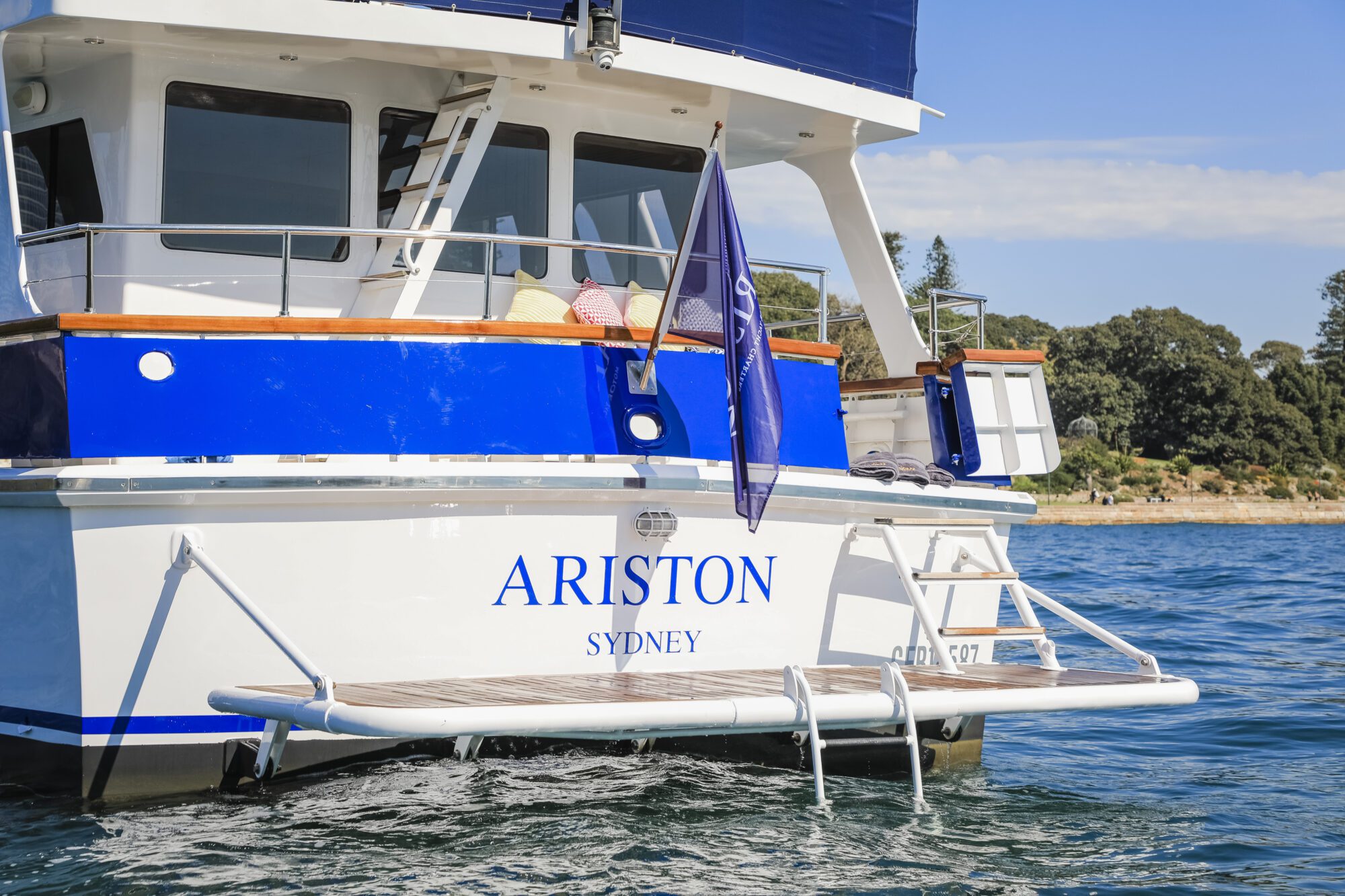 A blue and white boat named "ARISTON SYDNEY" anchored on clear blue water, with a swim platform and a flag at the stern. Cushions are visible on the deck, with trees and shoreline in the background.