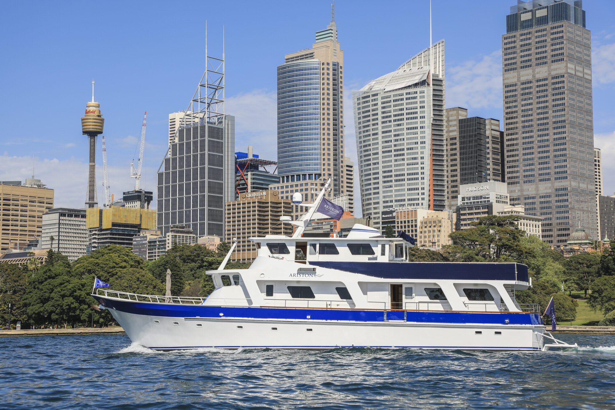 A white yacht with a blue stripe sails on the water, with a city skyline of modern high-rise buildings and a communications tower in the background, under a clear blue sky.