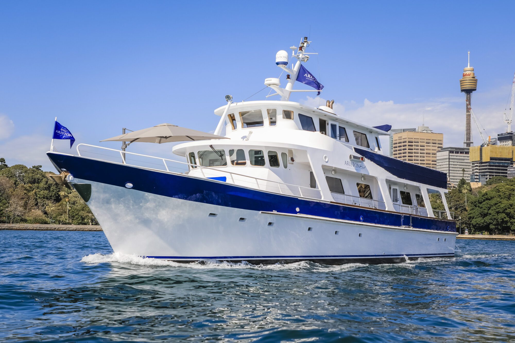 A large white and blue yacht cruises on the water near a city skyline, with a clear blue sky above and the Sydney Tower visible in the background.