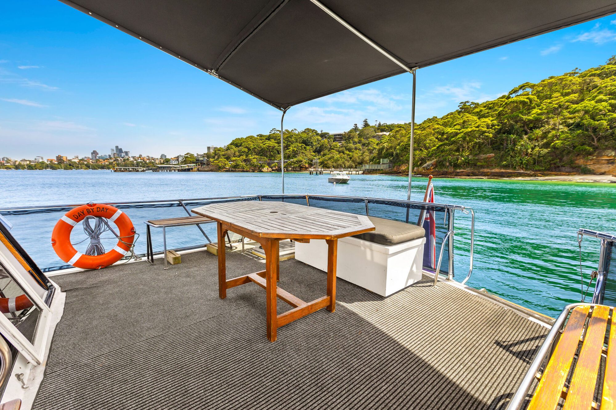 Covered deck area on a boat with a wooden table, bench seating, and a life buoy overlooking calm blue water and a green, tree-lined shoreline under a clear sky.
