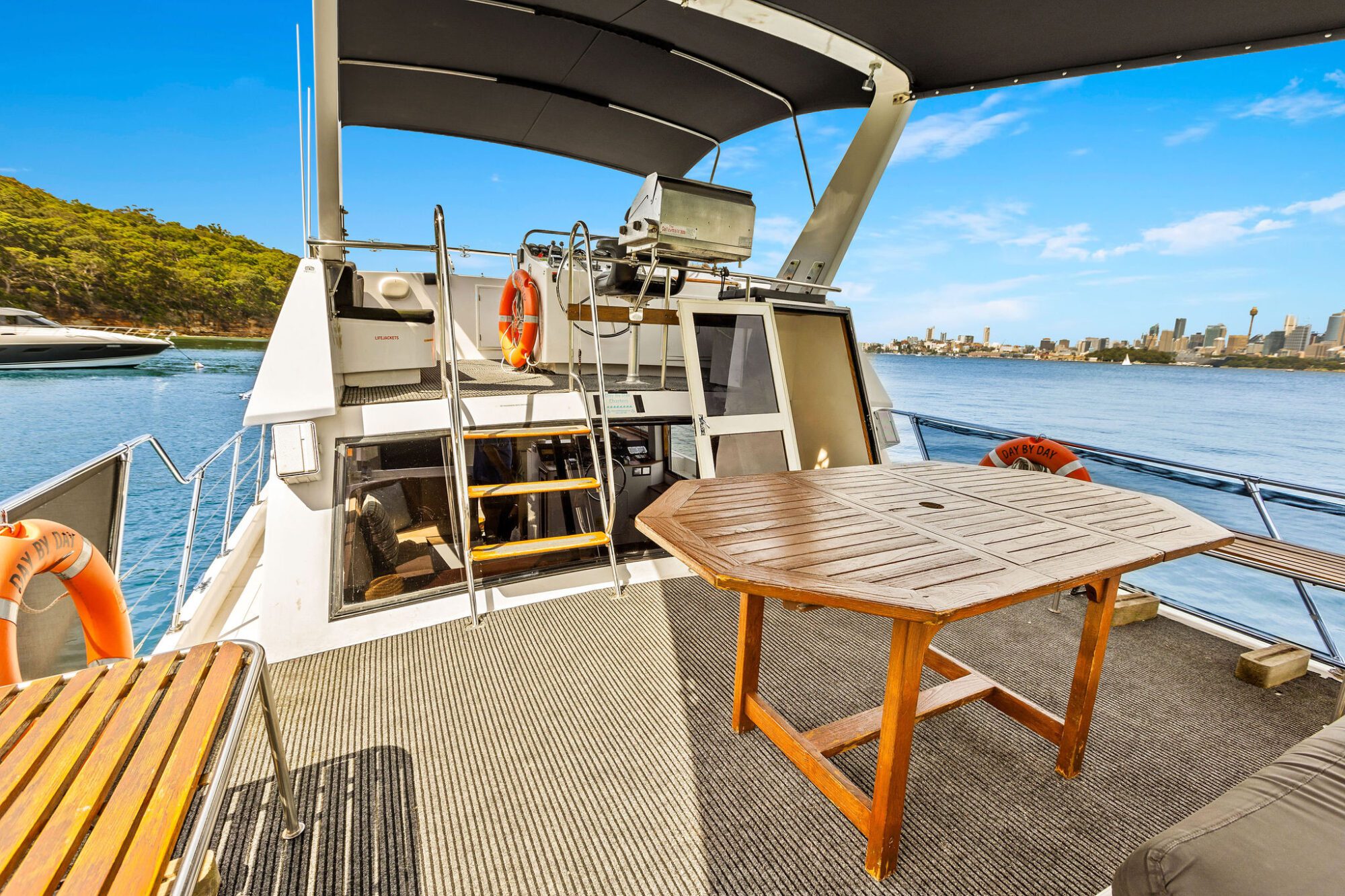 View from a boat’s deck with a wooden table, life rings, and stairs leading up to a covered upper area. The boat is on the water, with trees and city skyline visible in the background under a blue sky.