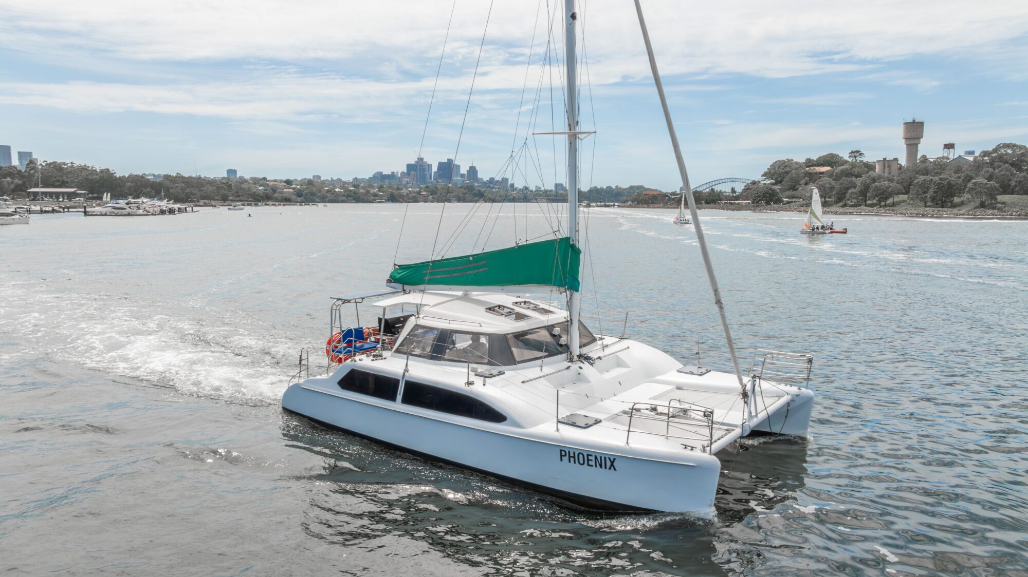 A white catamaran named "PHOENIX" sails on a calm body of water with a green canopy and city skyline in the background under a partly cloudy sky.
