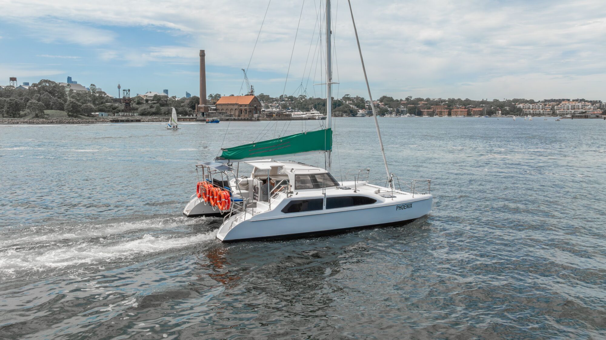 A white catamaran with a green canopy sails on a body of water, leaving a small wake behind. Life rings hang on the side, and buildings and trees are visible on the distant shore under a partly cloudy sky.