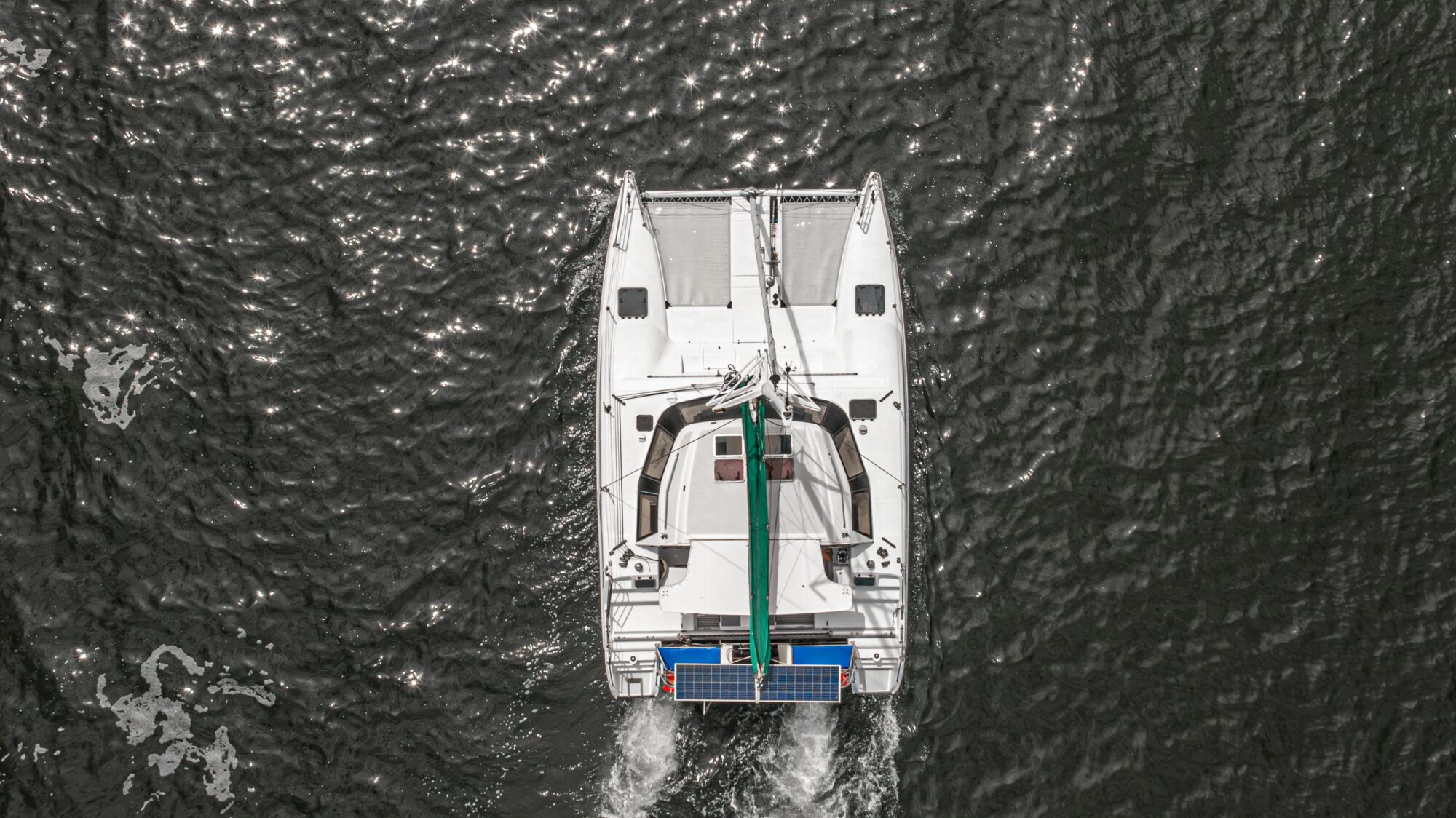 Aerial view of a white catamaran with solar panels on the stern, sailing on dark, rippling water, sunlight reflecting on the waves.
