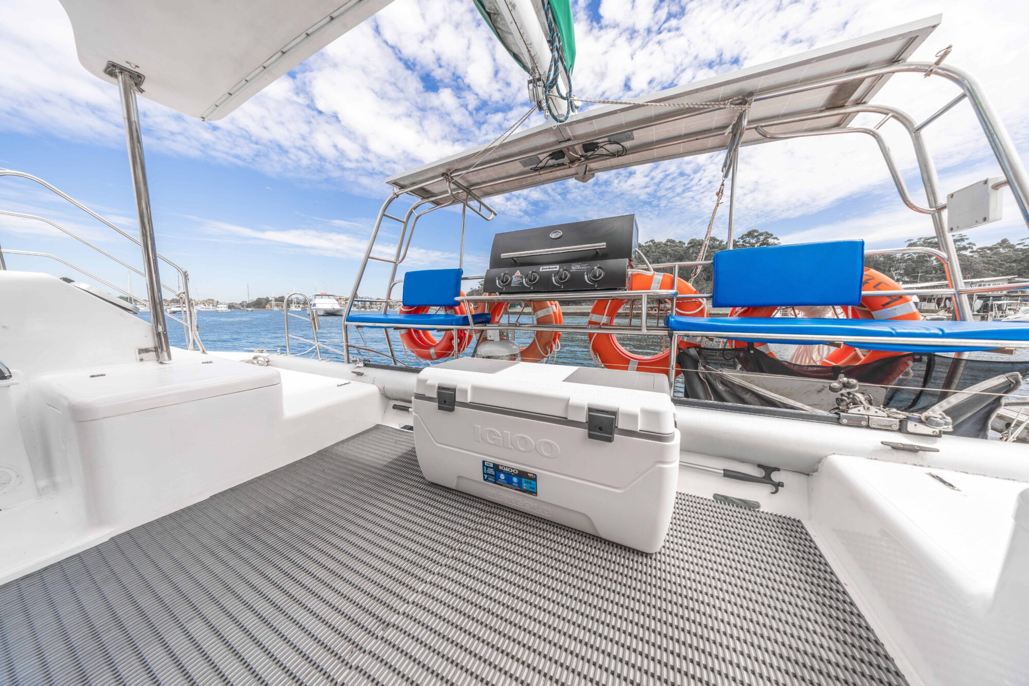 A white Igloo cooler sits on the deck of a boat near a stainless steel grill and orange life rings with blue holders, with a view of the water and sky in the background.