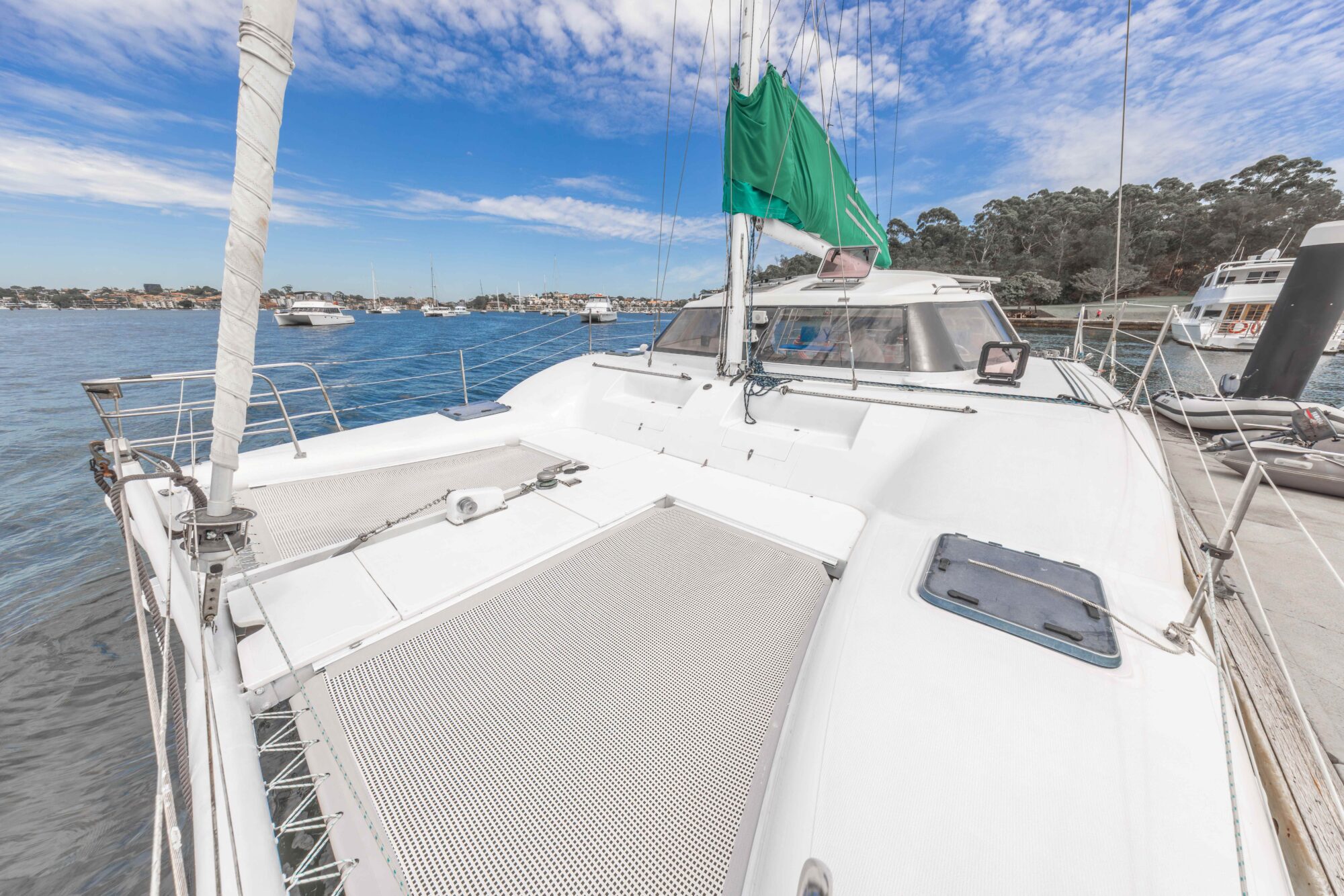 View from the deck of a white catamaran yacht docked at a marina, with calm blue water, anchored boats, and trees visible in the background under a partly cloudy sky.