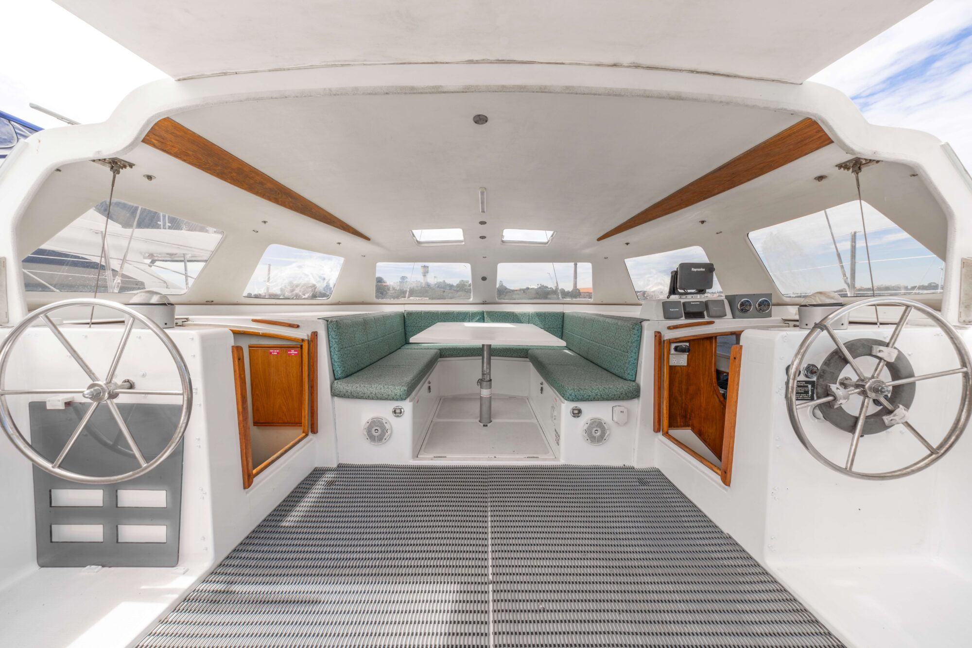 The image shows the interior of a catamaran cockpit with two steering wheels, green cushioned bench seating around a white table, wooden cabinets, and navigation equipment, all under a white ceiling with skylights.