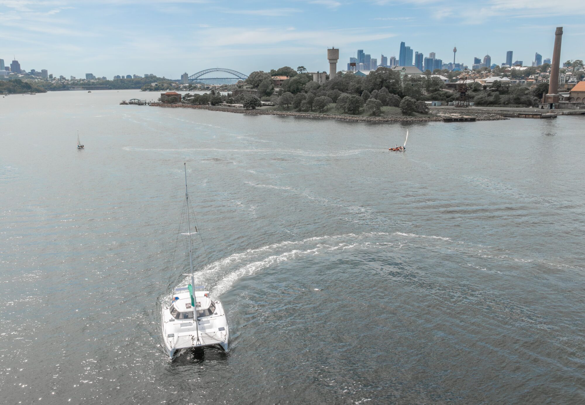 A white sailboat moves through a wide river with a city skyline, Sydney Harbour Bridge, and waterfront buildings visible in the background under a blue sky.
