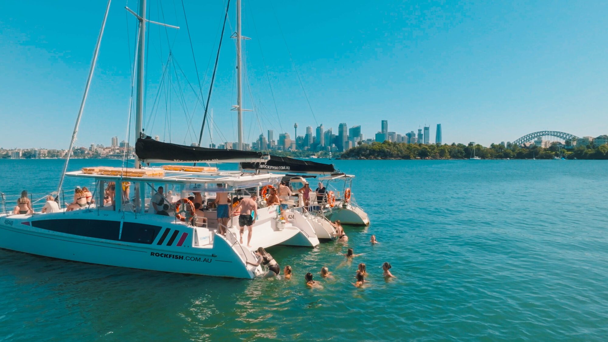 Two catamarans with people partying and swimming are anchored in blue water, with a city skyline and Sydney’s Harbour Bridge visible in the background under a clear, sunny sky.