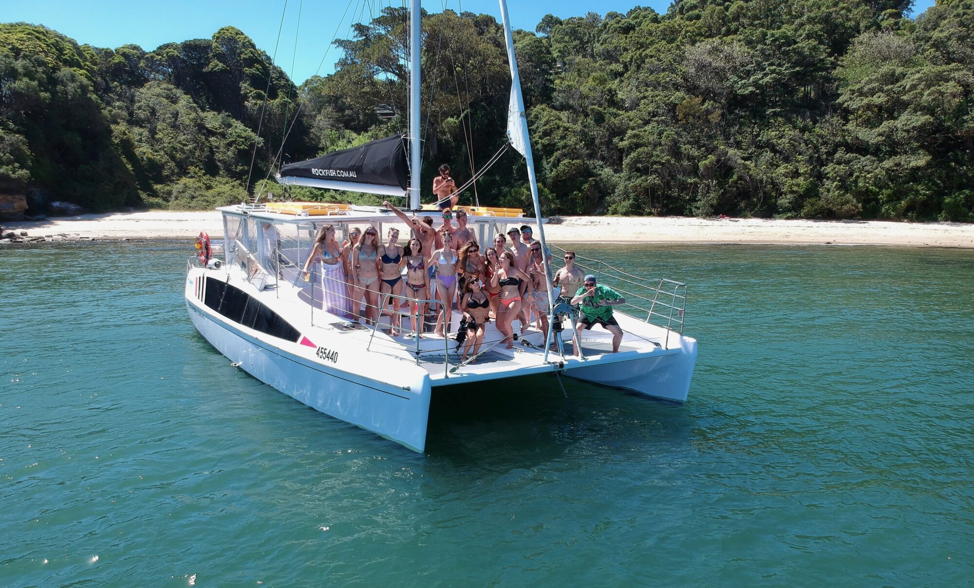 A group of people in swimwear stand and pose on the deck of a white catamaran anchored in clear blue-green water near a sandy beach with lush green trees in the background.