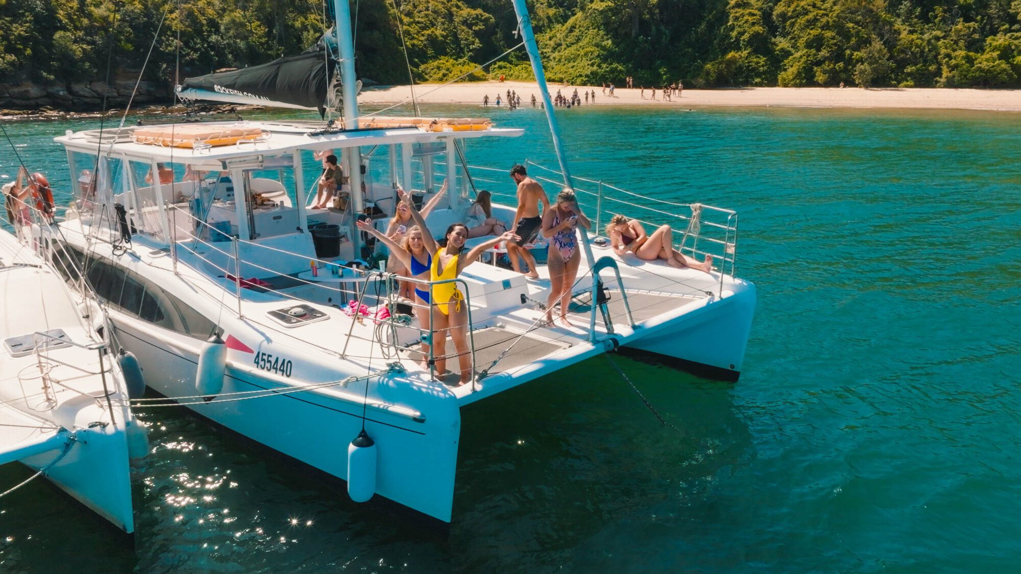 A group of people in swimsuits stand and relax on a white catamaran anchored near a tropical beach, with clear turquoise water and lush green trees in the background.