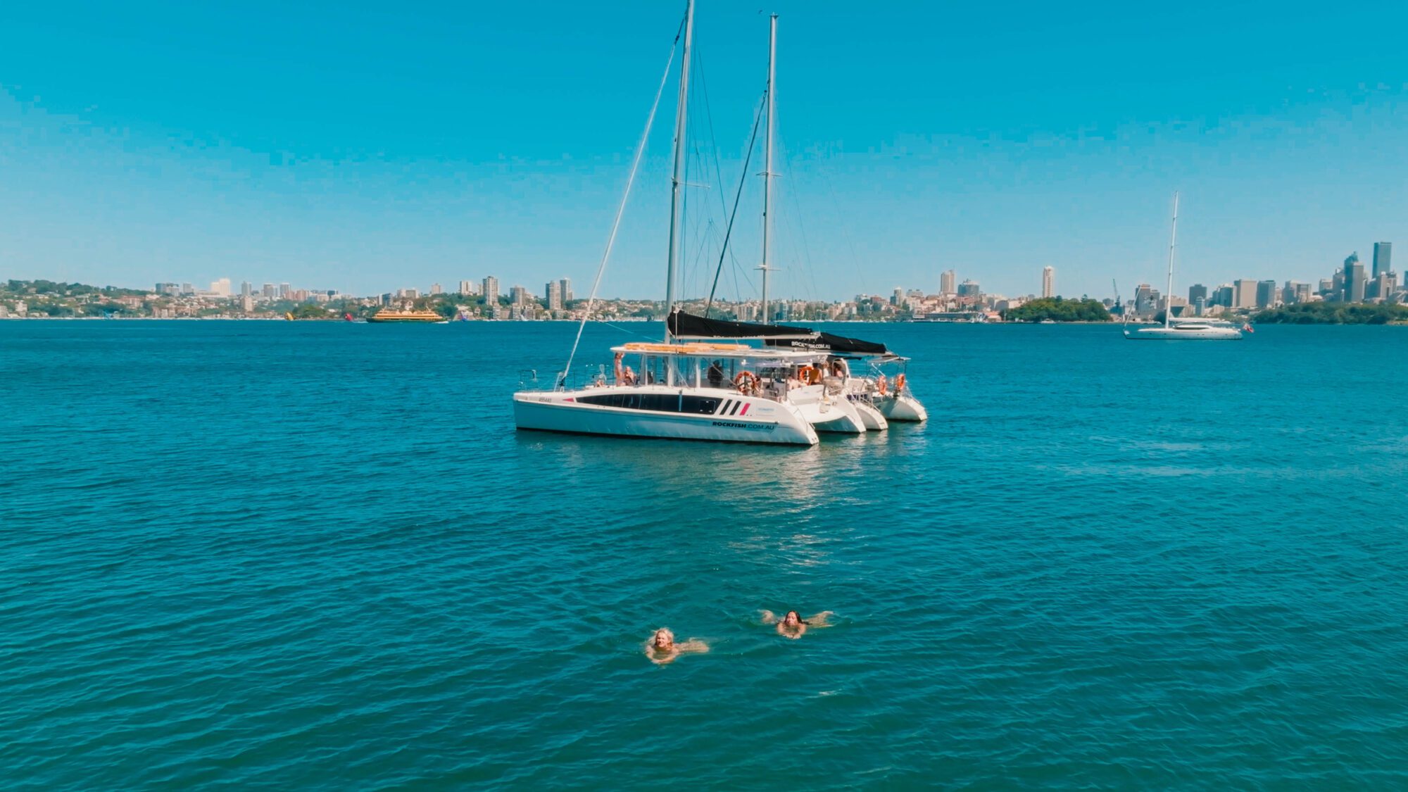 A catamaran with people onboard floats on blue water near a city skyline, while three people swim nearby under a clear, sunny sky.