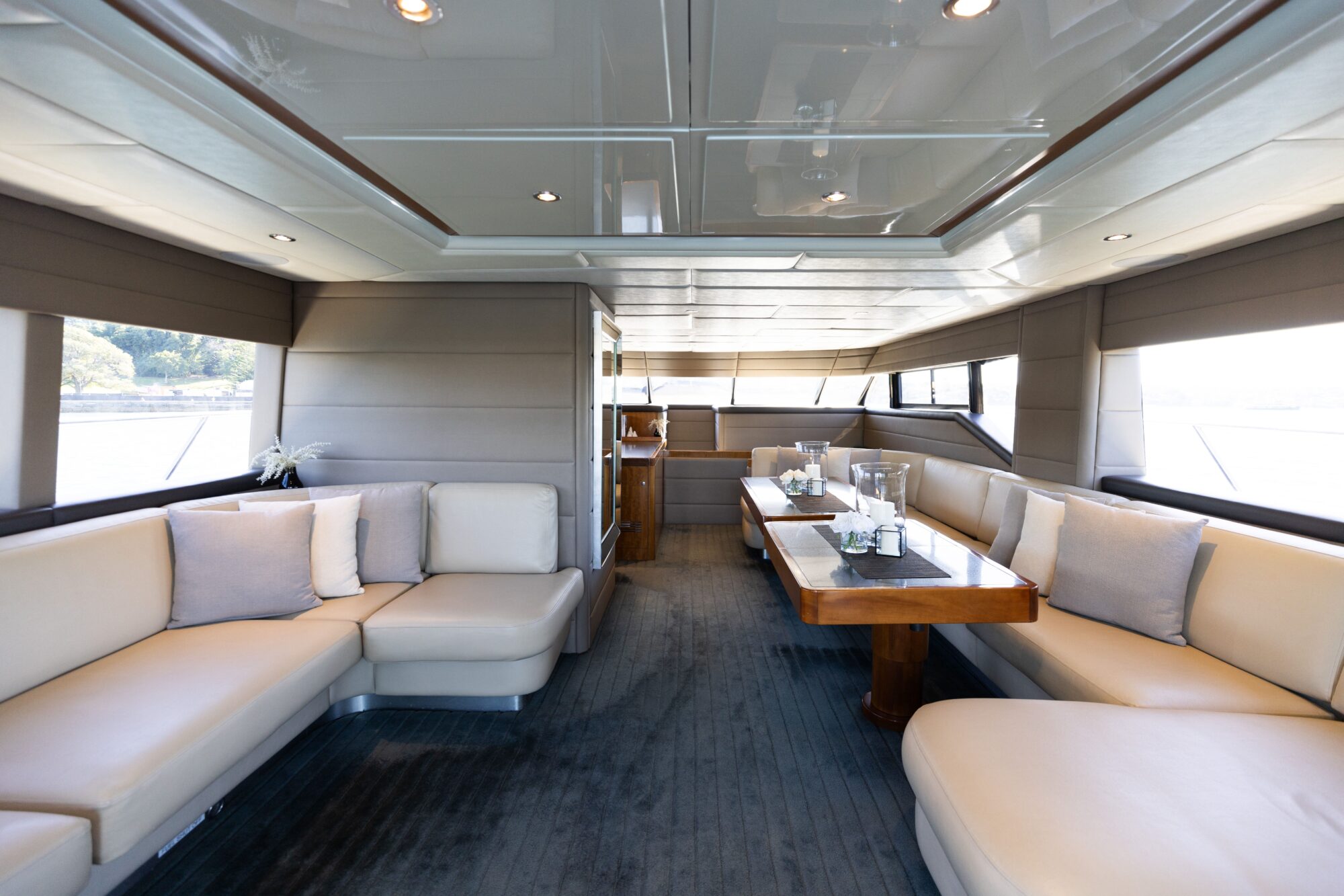 The image shows the interior of a luxury yacht with cream-colored leather seating, wooden tables set with glassware, large windows, and modern decor offering views of the water outside.