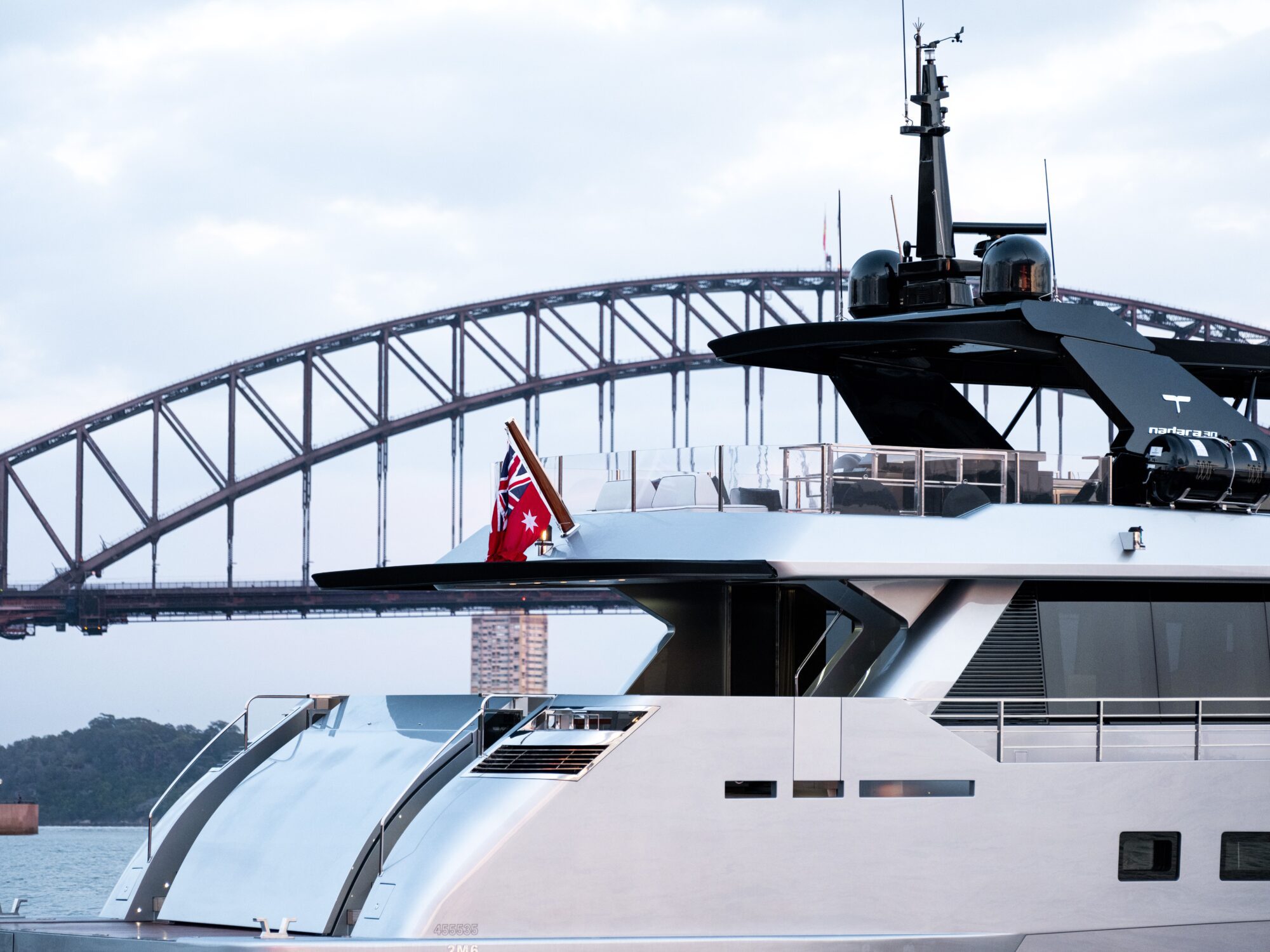 A luxury yacht with a red ensign flag is docked in front of the Sydney Harbour Bridge, under a cloudy sky.