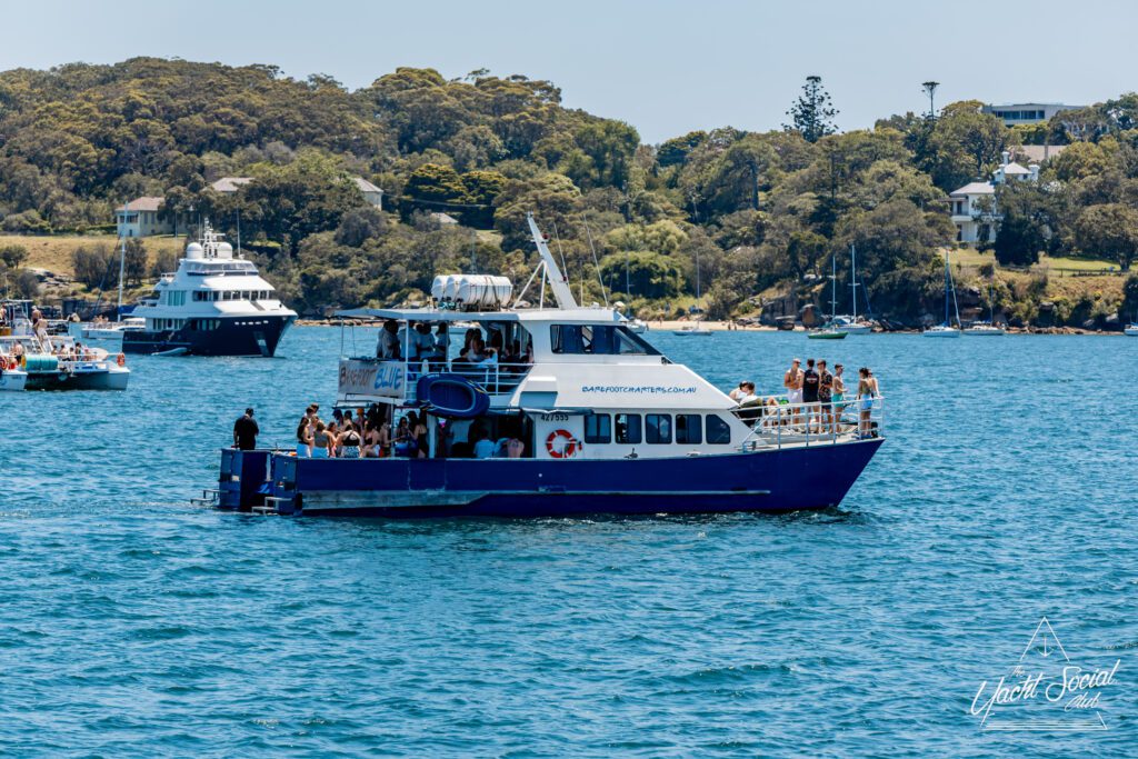 A blue and white party boat filled with people is cruising on a sunny day, surrounded by clear blue water and other boats, with green trees and houses visible on the shoreline in the background.