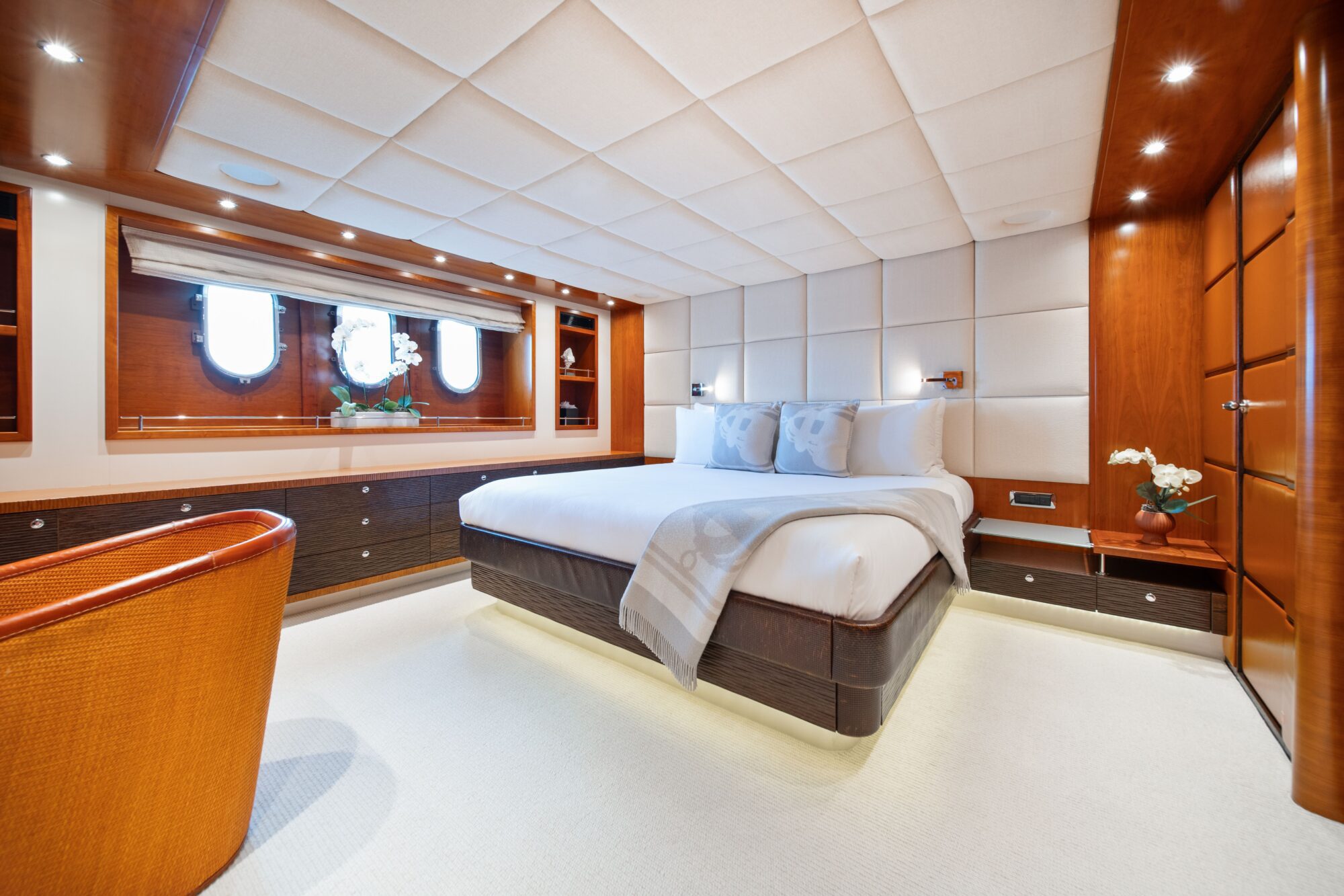 A luxurious yacht bedroom with a large bed, soft white bedding, wood paneling, padded walls, round windows, built-in shelving, a cozy chair, and warm ambient lighting.