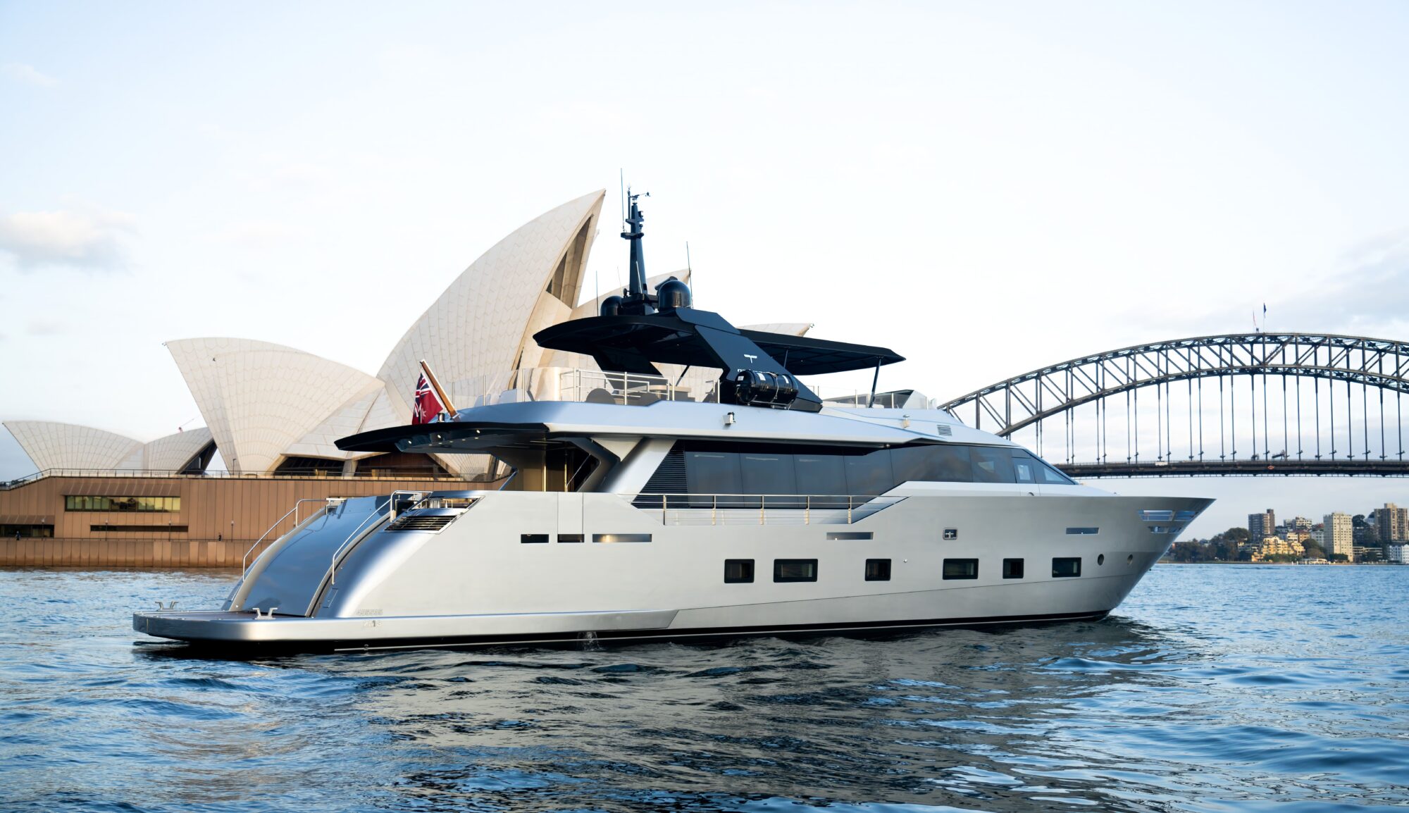 A modern luxury yacht sails on the water with the Sydney Opera House and Sydney Harbour Bridge visible in the background on a clear day.