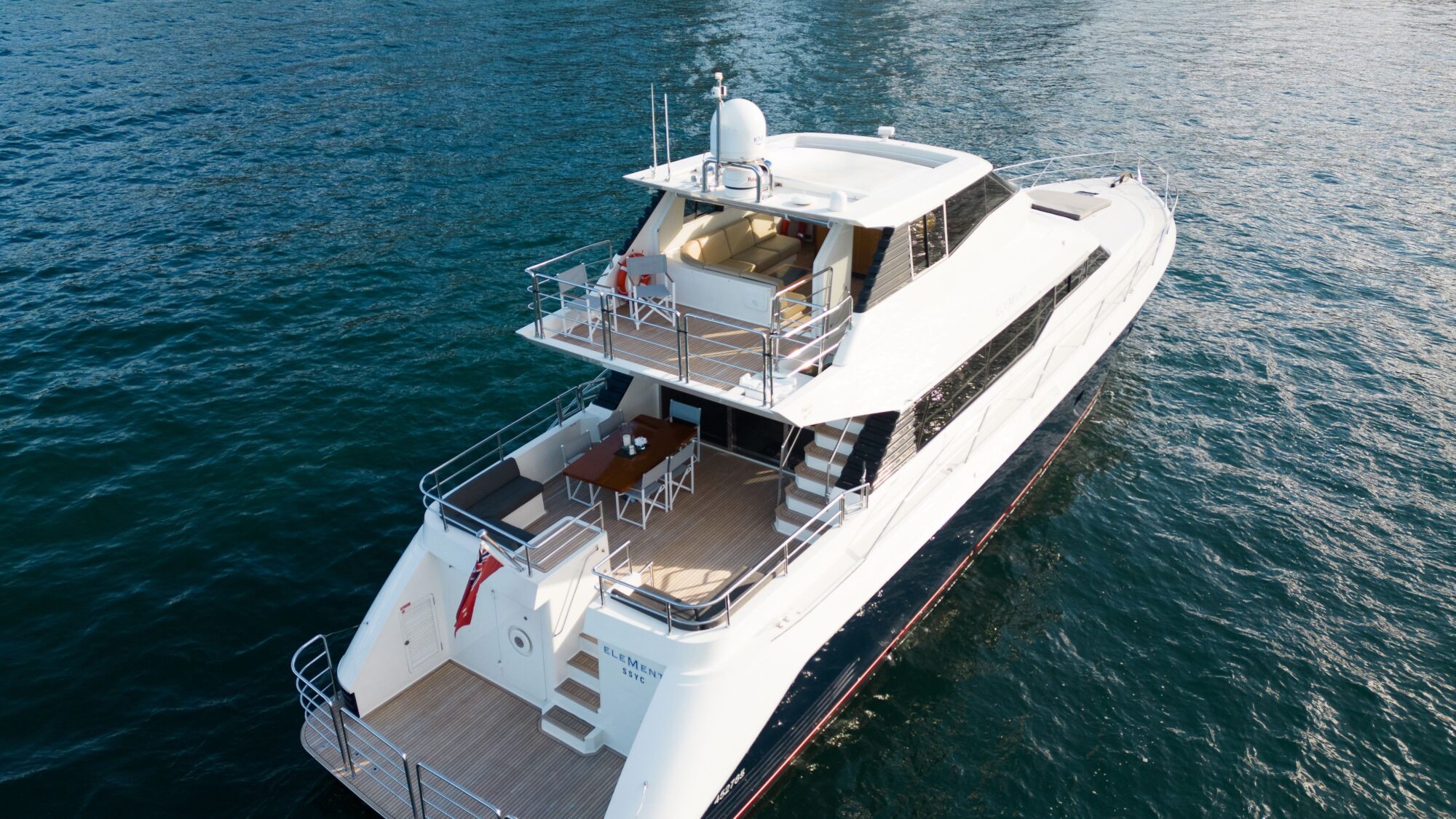 A white luxury yacht with two decks floats on calm blue water. The upper deck features a seating area, while the lower deck has a dining table and chairs, with railings surrounding both levels.