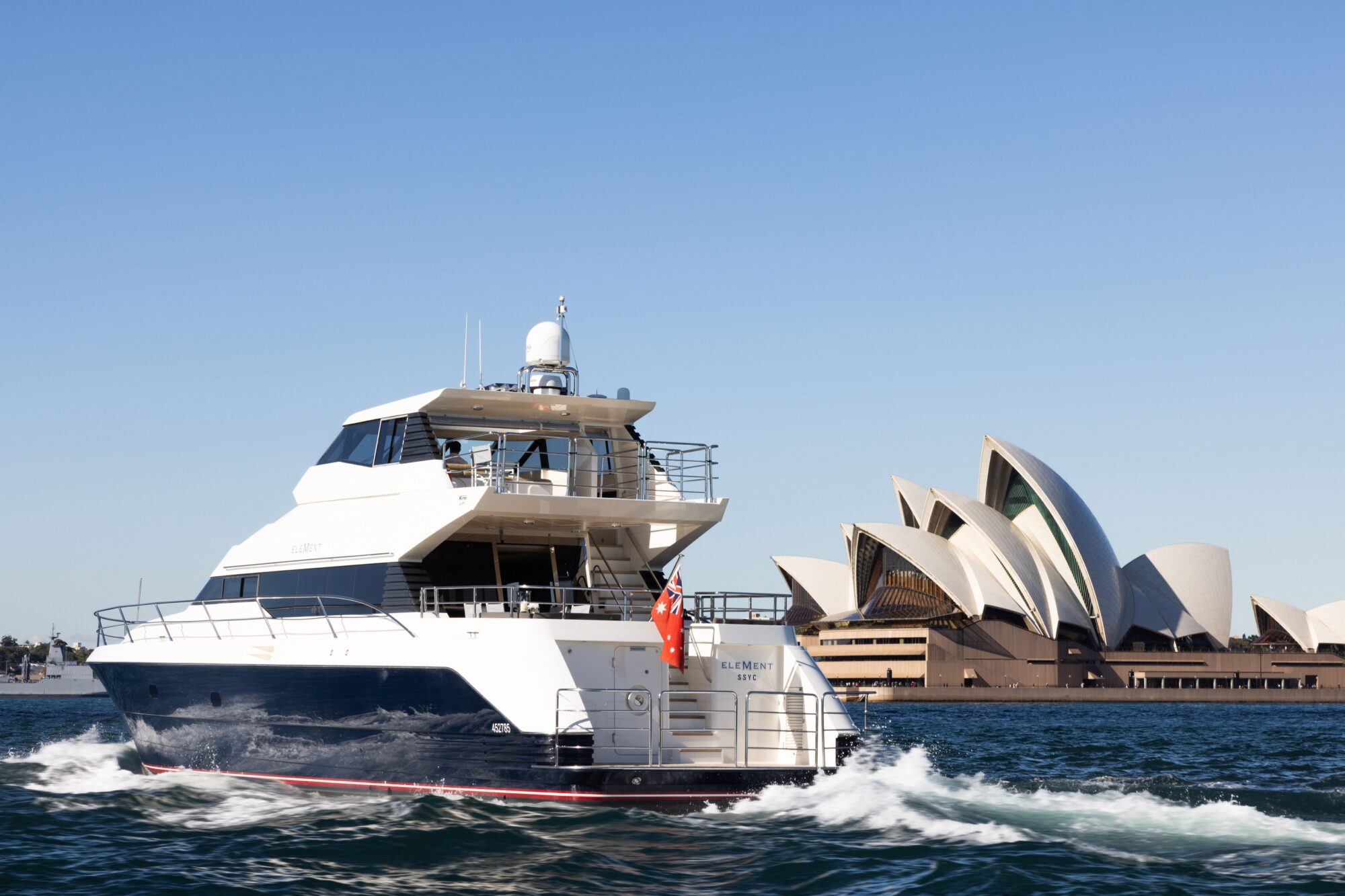 A luxury yacht sails on blue water near the Sydney Opera House in Australia, with clear skies overhead and waves trailing behind the boat.