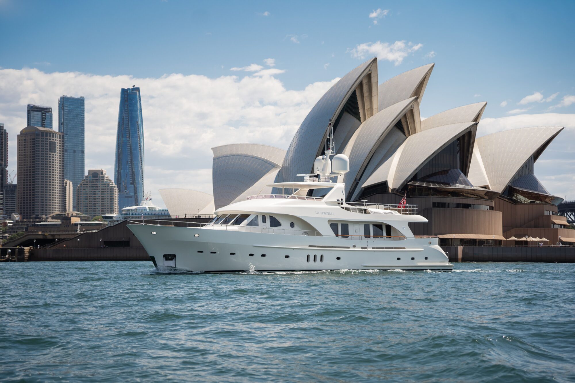 A white yacht sails on the water in front of the Sydney Opera House, with tall modern buildings in the background under a partly cloudy sky.