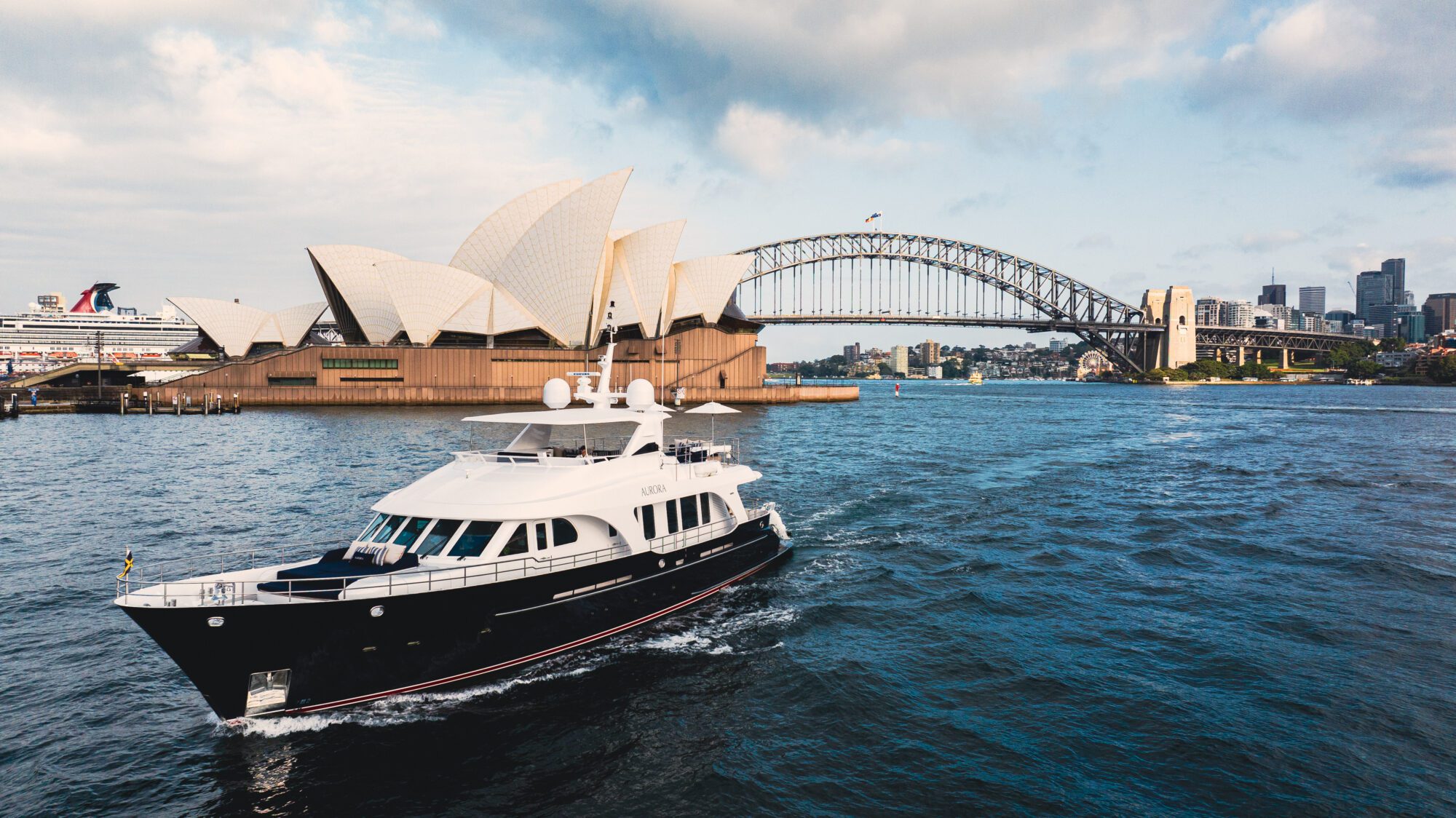 A luxury yacht sails on Sydney Harbour with the Sydney Opera House and Sydney Harbour Bridge visible in the background under a partly cloudy sky.