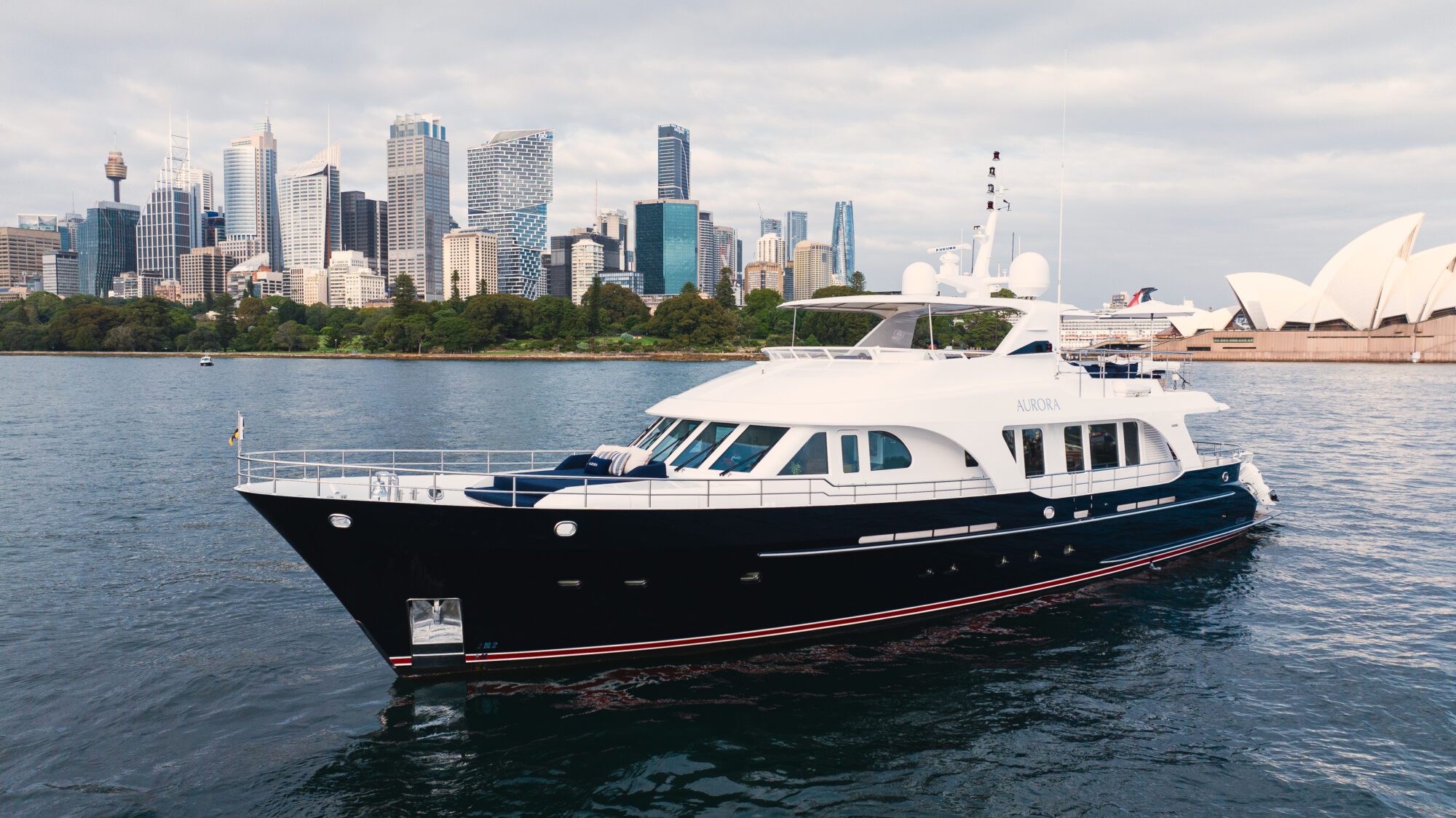 A large luxury yacht sails on the water with a modern city skyline and the Sydney Opera House in the background under a cloudy sky.