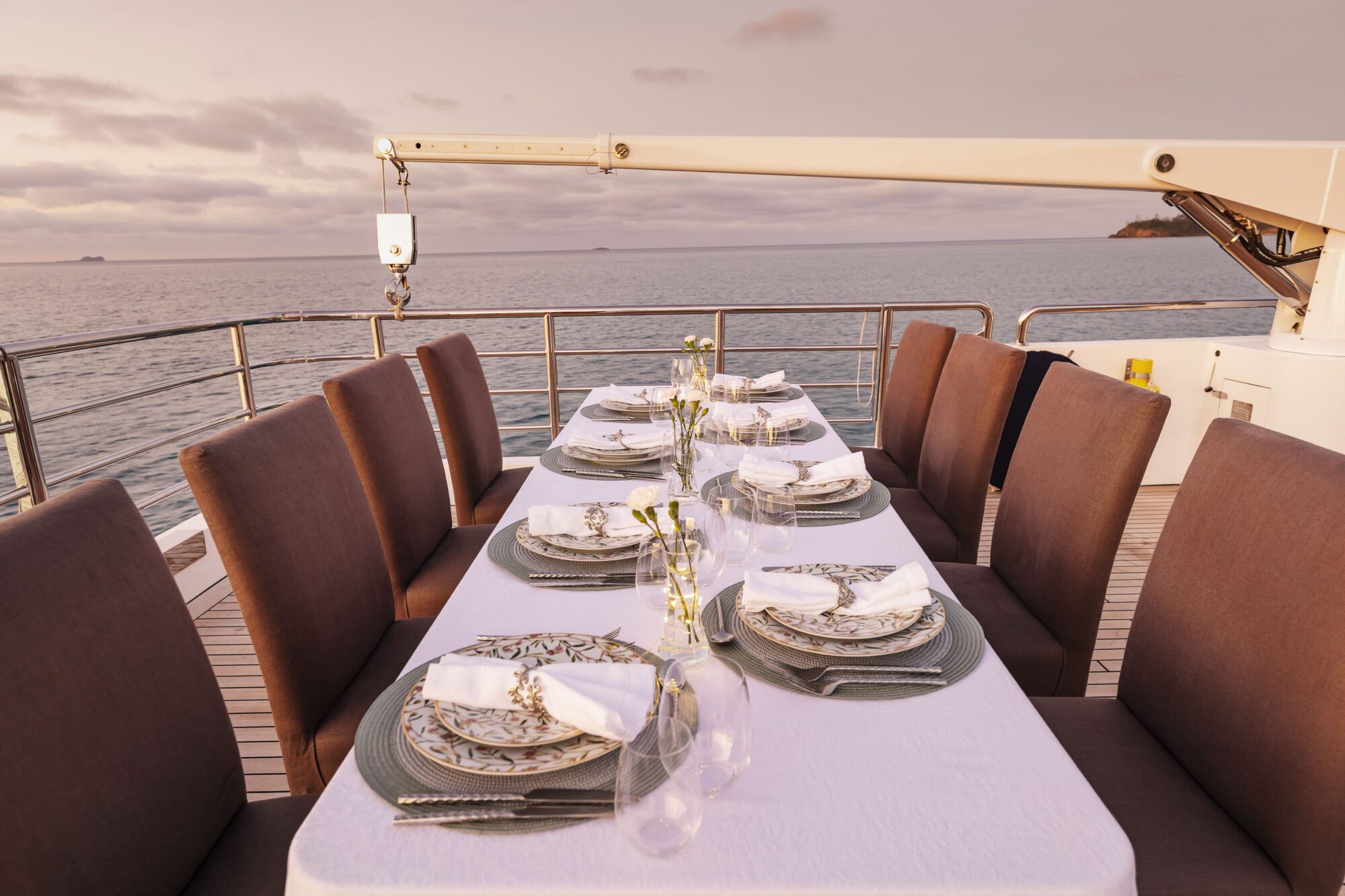 Elegant outdoor dining table set for a meal on the deck of a yacht at sunset, with patterned plates, napkins, glasses, and small flower vases, overlooking calm ocean waters.