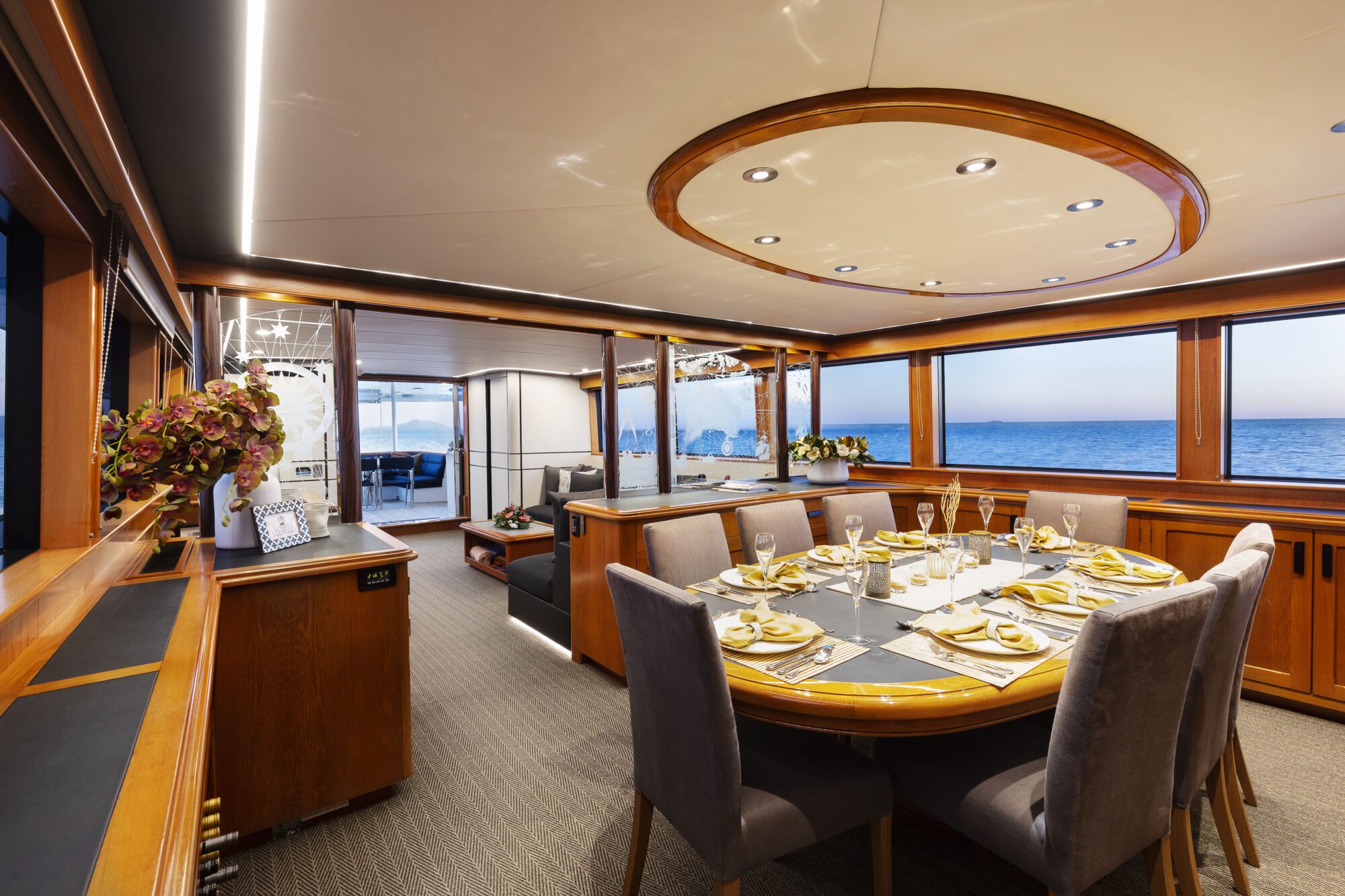 A luxurious yacht dining area features an elegant set table with gold-accented plates and glassware, surrounded by gray chairs, large windows overlooking the ocean, and warm wooden finishes throughout the interior.