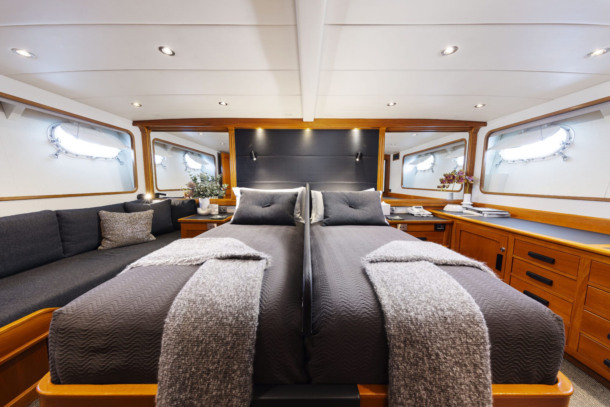 A luxurious yacht cabin with two single beds side by side, dark gray bedding, and gray throws. The space features wood paneling, a sofa, built-in dressers, mirrors, and windows letting in natural light.