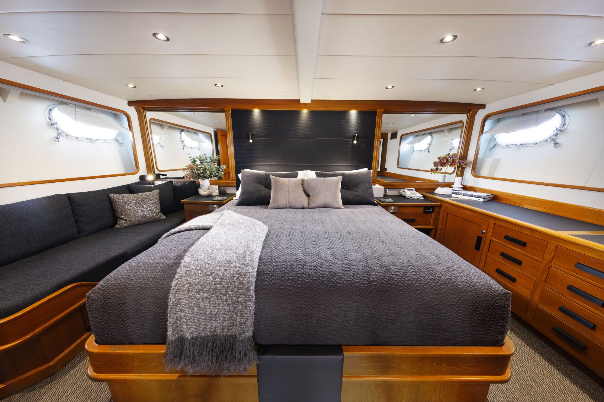 Luxury yacht bedroom with a large bed covered in gray bedding, flanked by wooden cabinets and windows. A gray throw and decorative pillows are on the bed, with a black sofa and flowers on the left side.