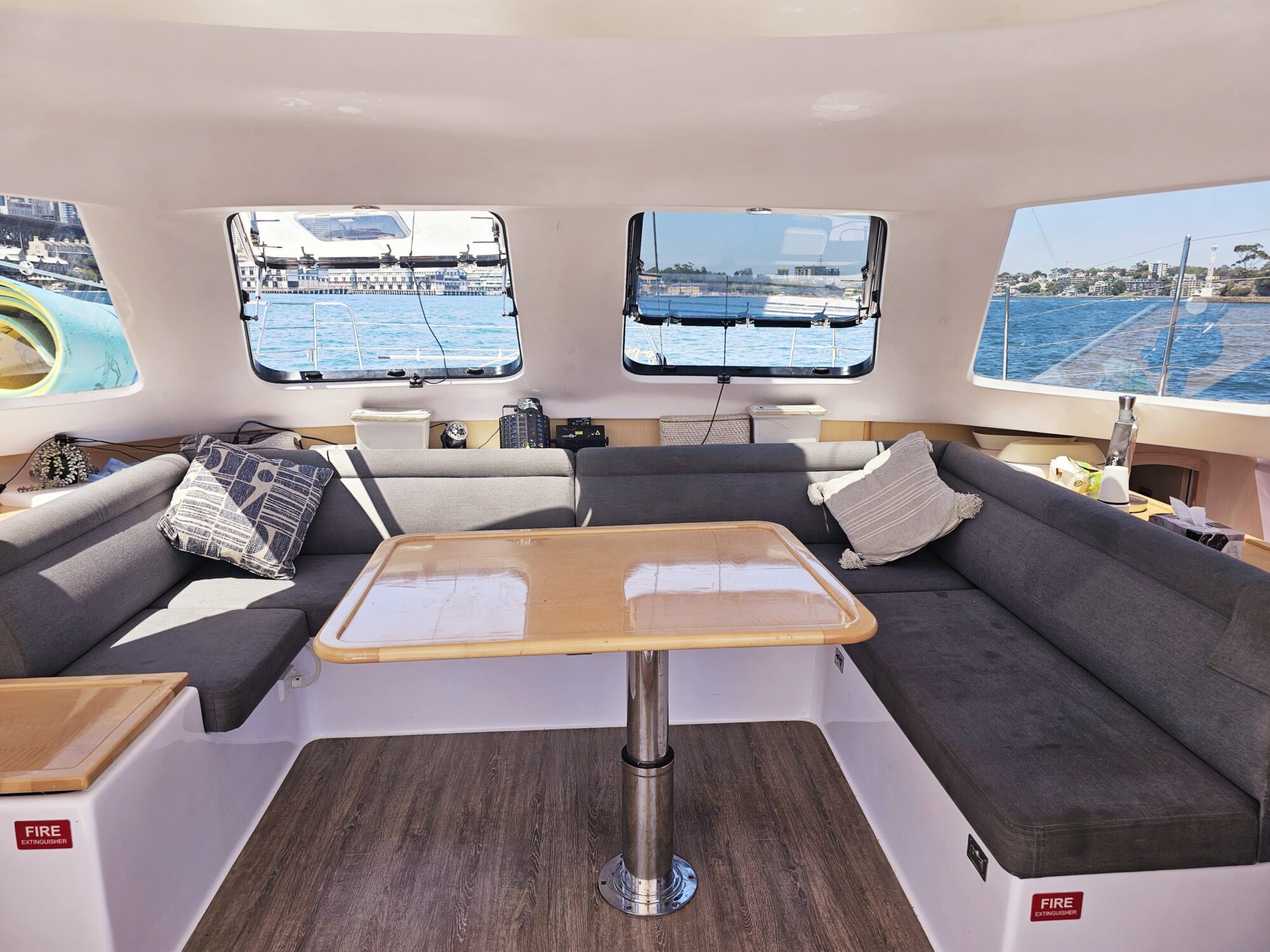 The interior of a boat with gray cushioned seating, a wooden table, large windows showing a blue sea and shore, and sunlight streaming in. Pillows are on the seats, and fire safety signs are visible.