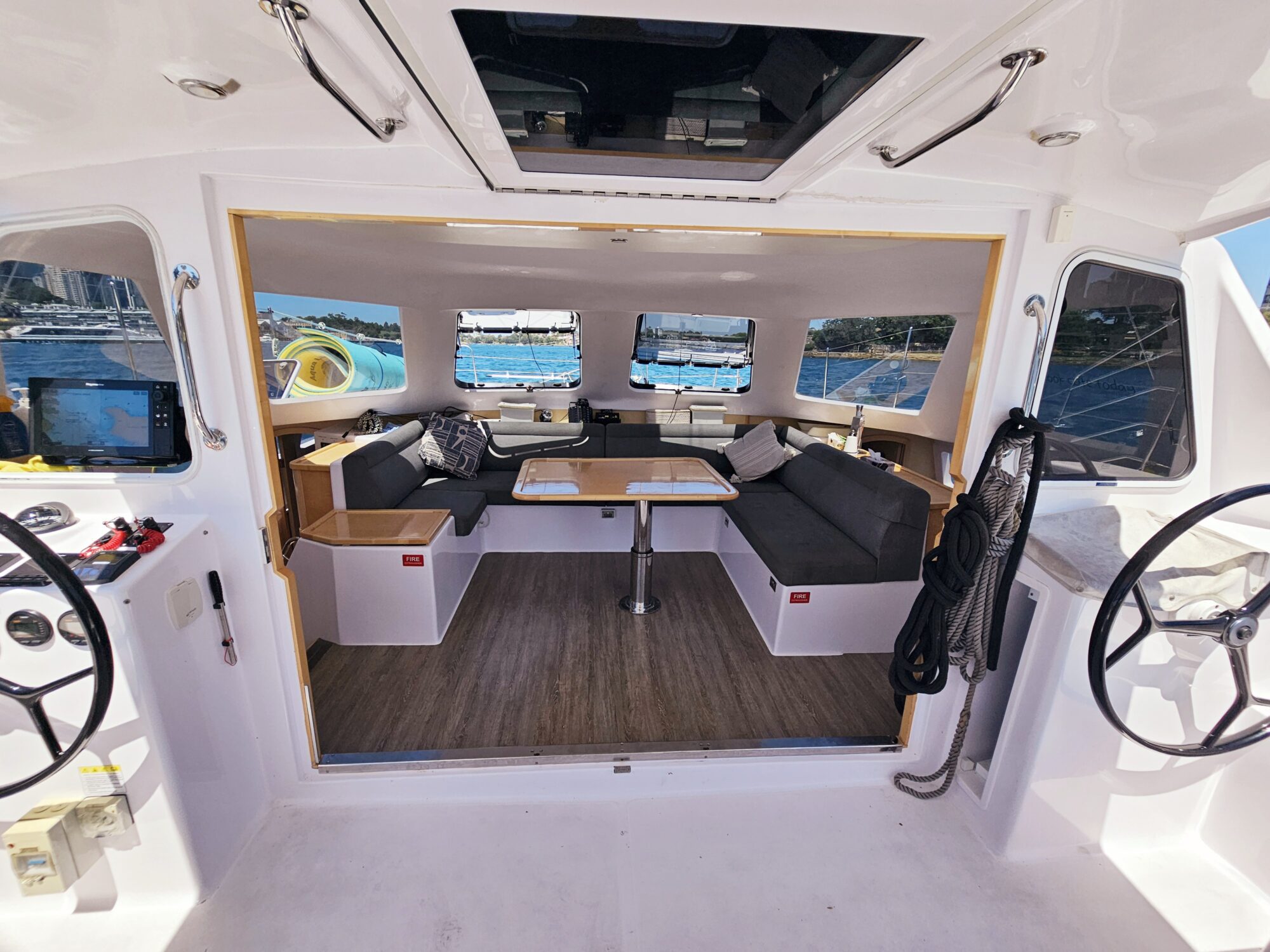 View of a catamaran's spacious interior lounge with gray seating, a wooden table, large windows showing a marina and water outside, and steering wheels on either side of the entrance.