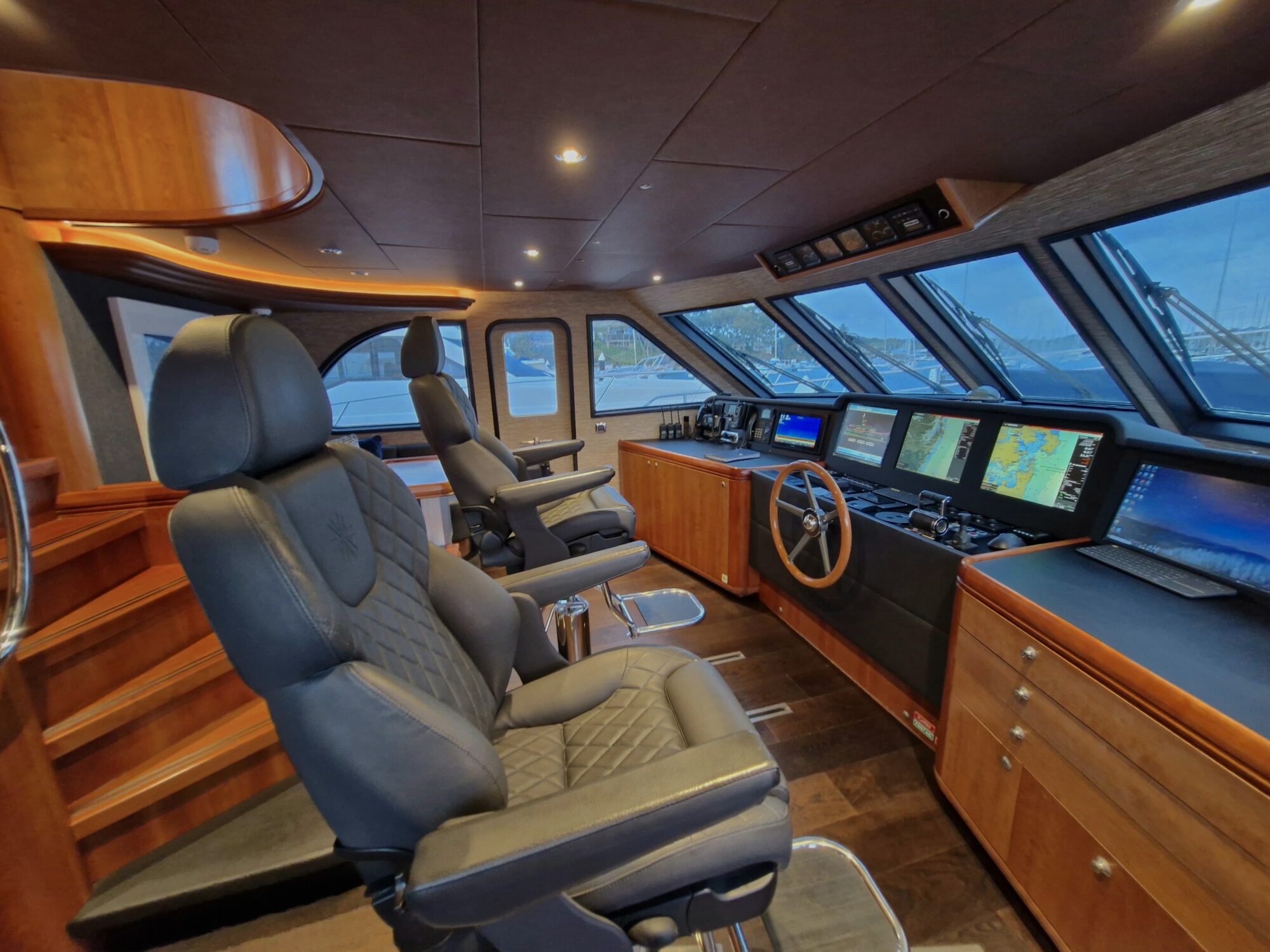 Modern yacht cockpit with two black leather captain’s chairs, multiple navigation screens, wooden cabinets, and large windows offering a view of the marina outside.