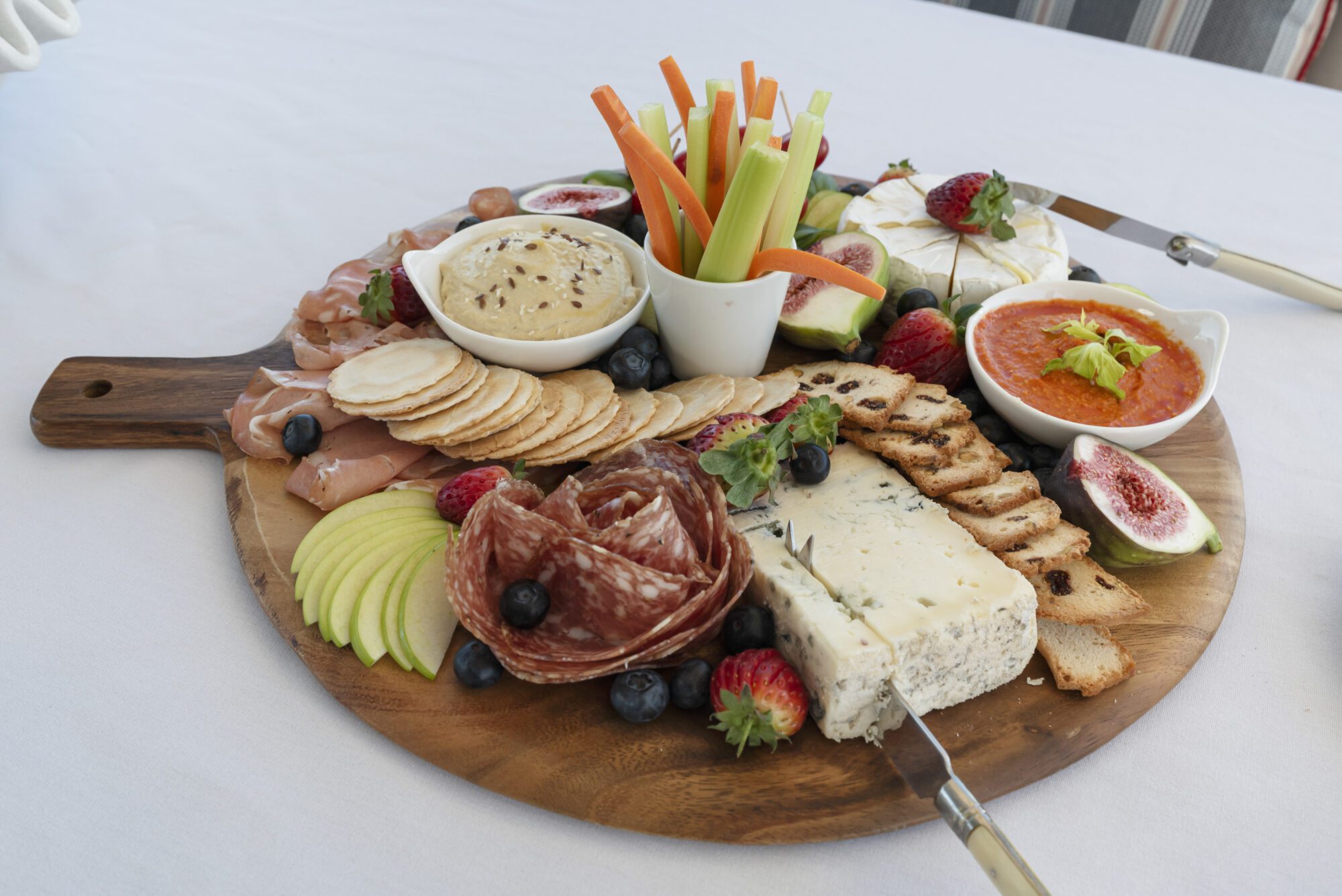 A wooden platter with assorted cheeses, crackers, salami, apple slices, fresh berries, figs, vegetable sticks, a small bowl of hummus, a bowl of red dip, and slices of bread on a white tablecloth.