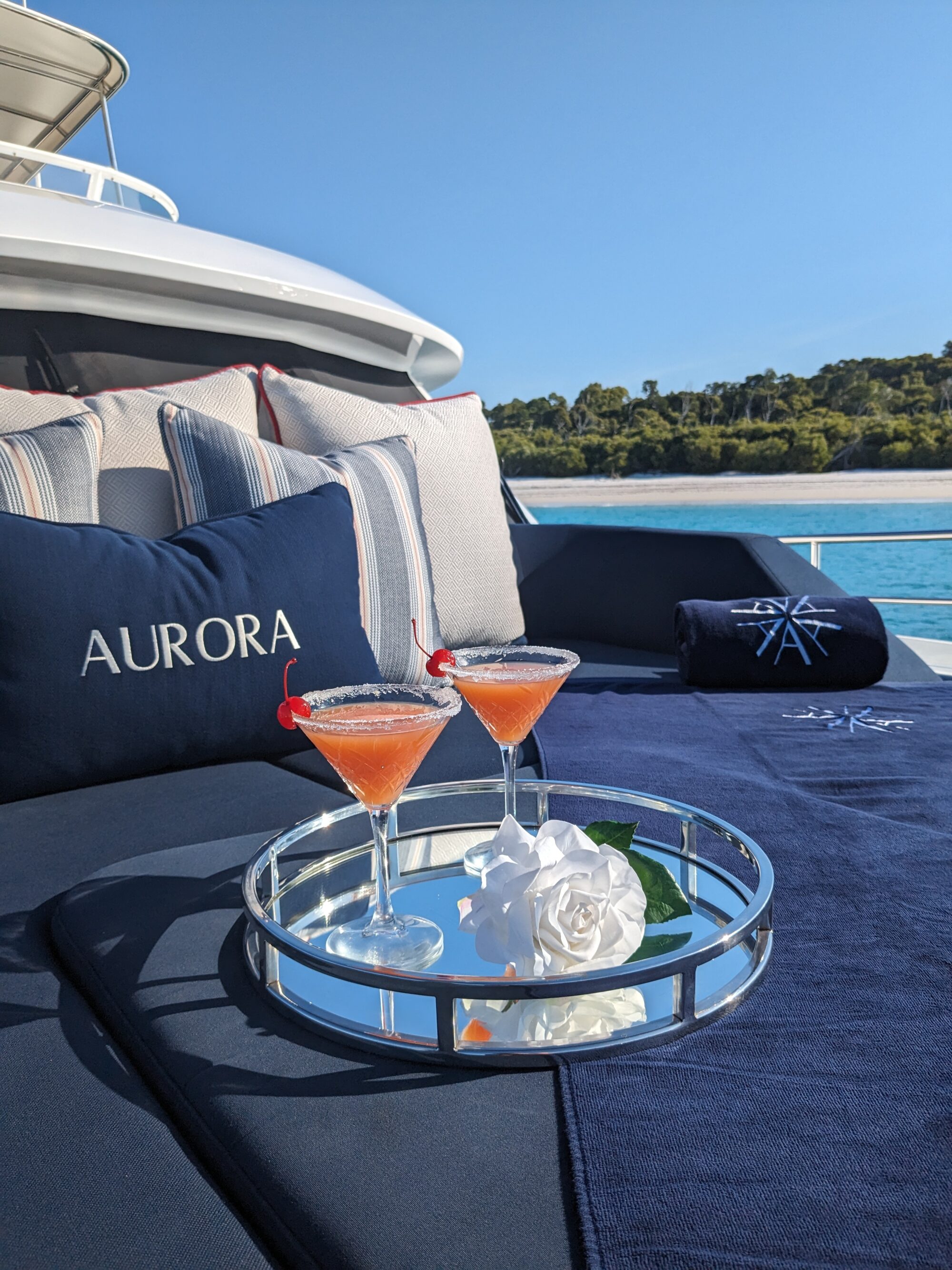 Two pink cocktails with cherries sit on a silver tray alongside a white rose on a yacht’s outdoor lounge. Blue pillows and towels with "Aurora" embroidered are visible, with a view of the sea and trees in the background.