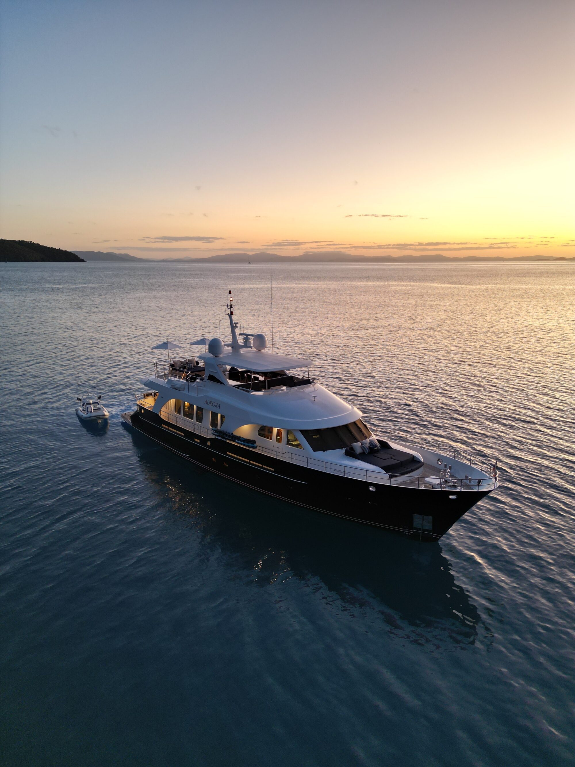 A luxury yacht is anchored on calm blue water at sunset, with a small boat floating nearby. The sky is warm with golden hues, and distant land is visible on the horizon.