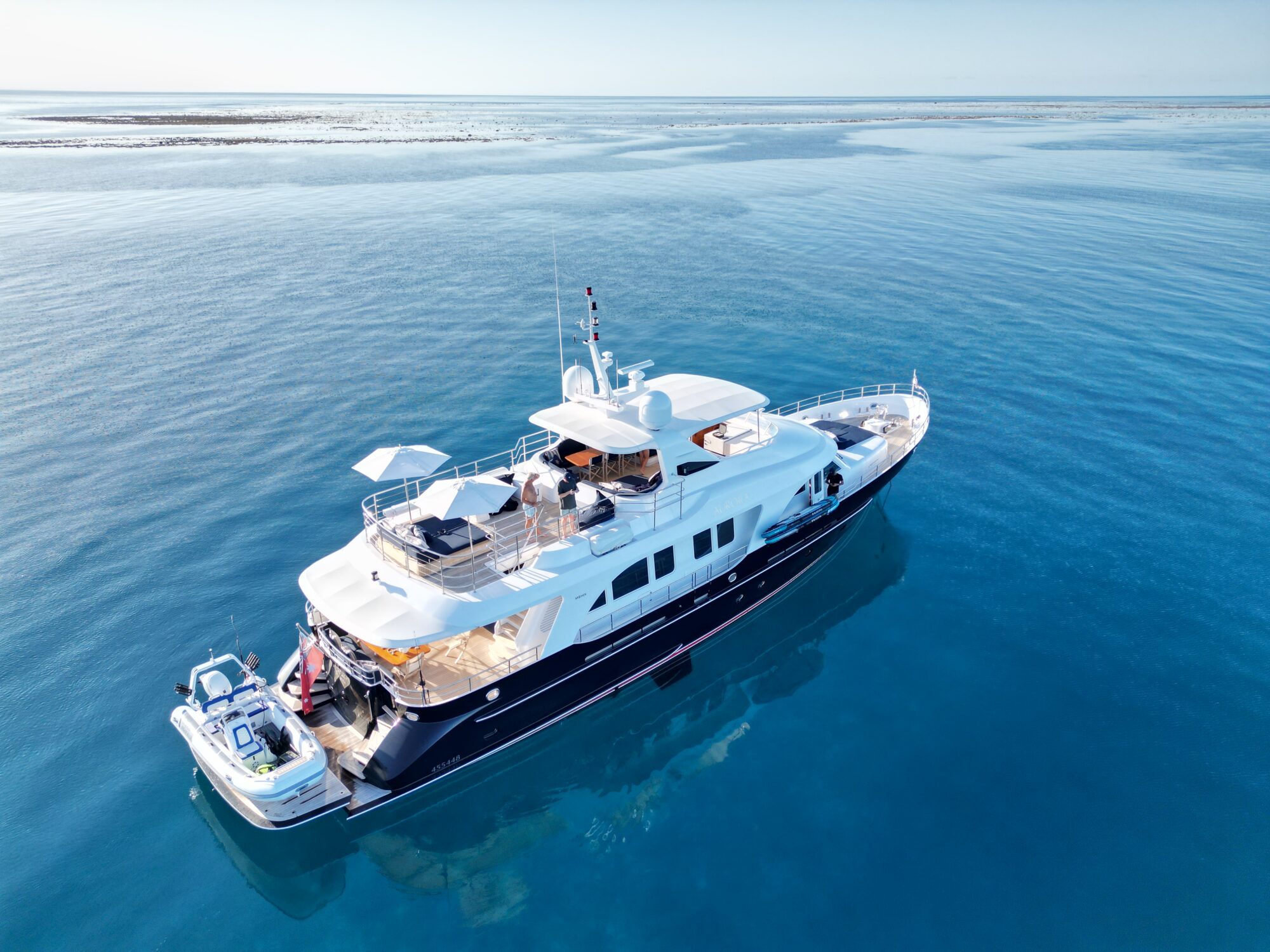 A luxury yacht with people relaxing on deck floats on calm, clear blue water under a sunny sky, with a small boat attached at the back and distant land barely visible on the horizon.