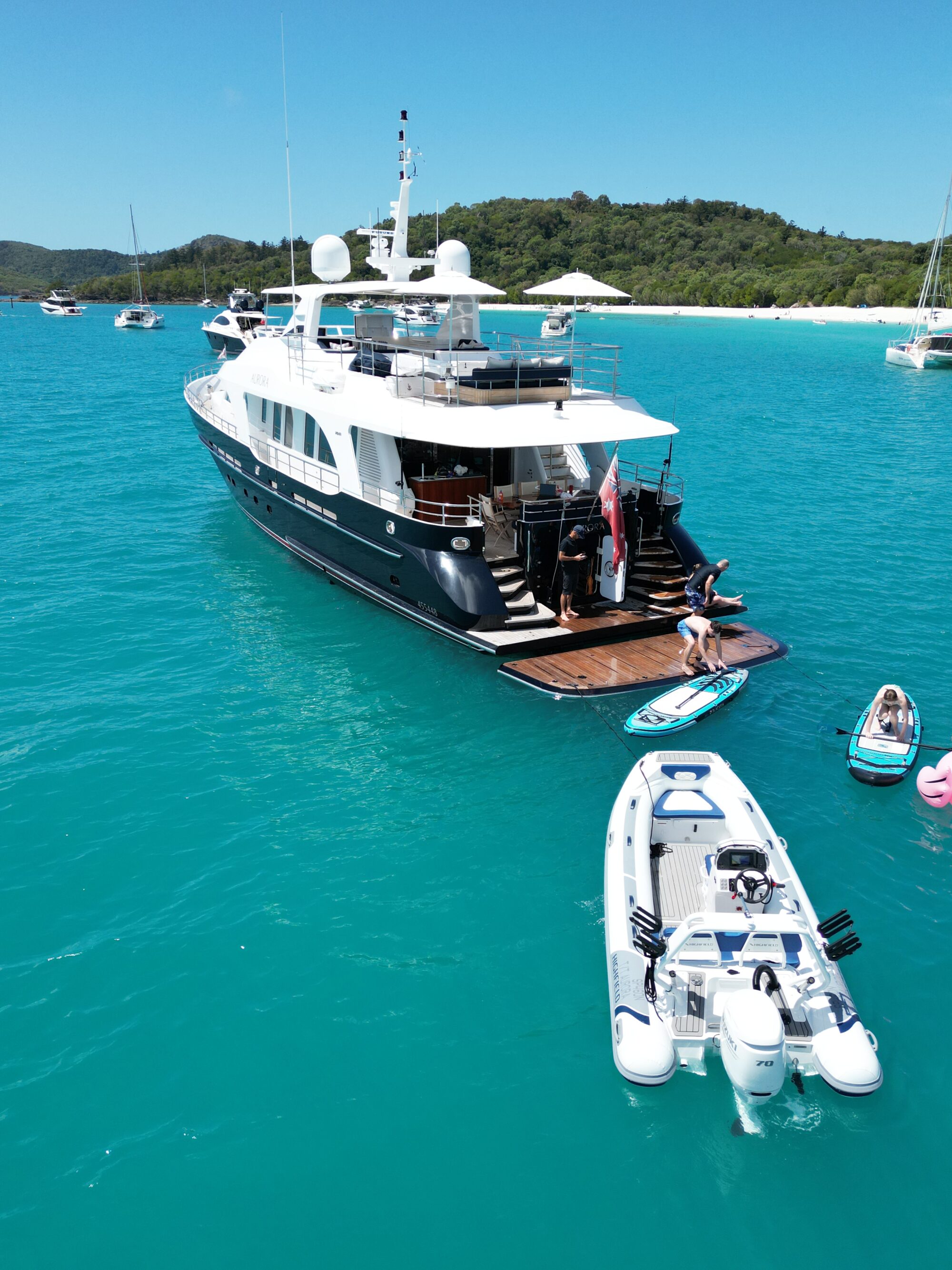 A luxury yacht anchored in turquoise water near a beach with lush green hills. People are near the yacht, with an inflatable boat and paddleboards nearby. Several other boats are visible in the background.