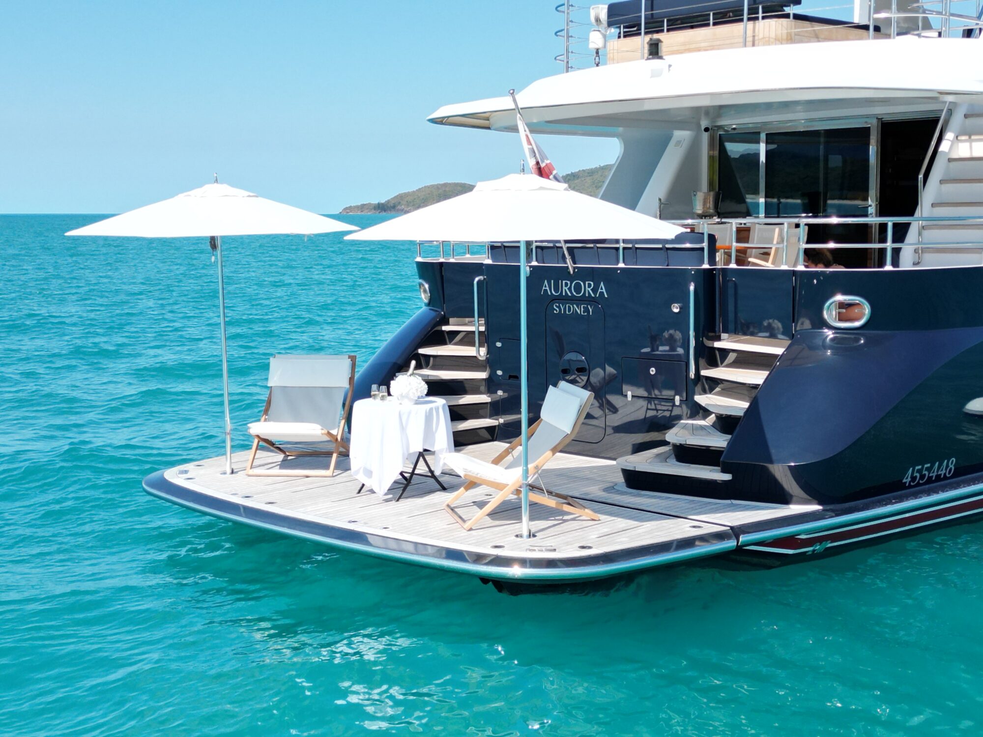 A luxury yacht named "Aurora Sydney" anchored in turquoise water, featuring a deck with two lounge chairs, a white table set with drinks, and two white umbrellas. Distant land is visible on the horizon.
