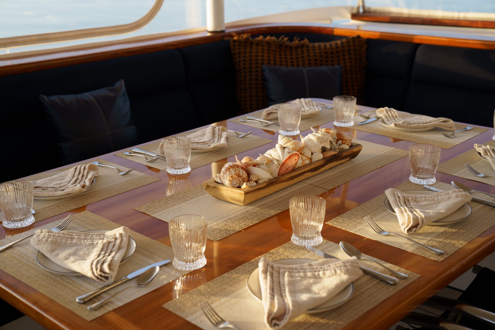 A polished wooden dining table set for six, featuring beige napkins, crystal glasses, cutlery, and a centerpiece platter with assorted seafood, on a boat with dark cushioned seating and sunlight streaming in.