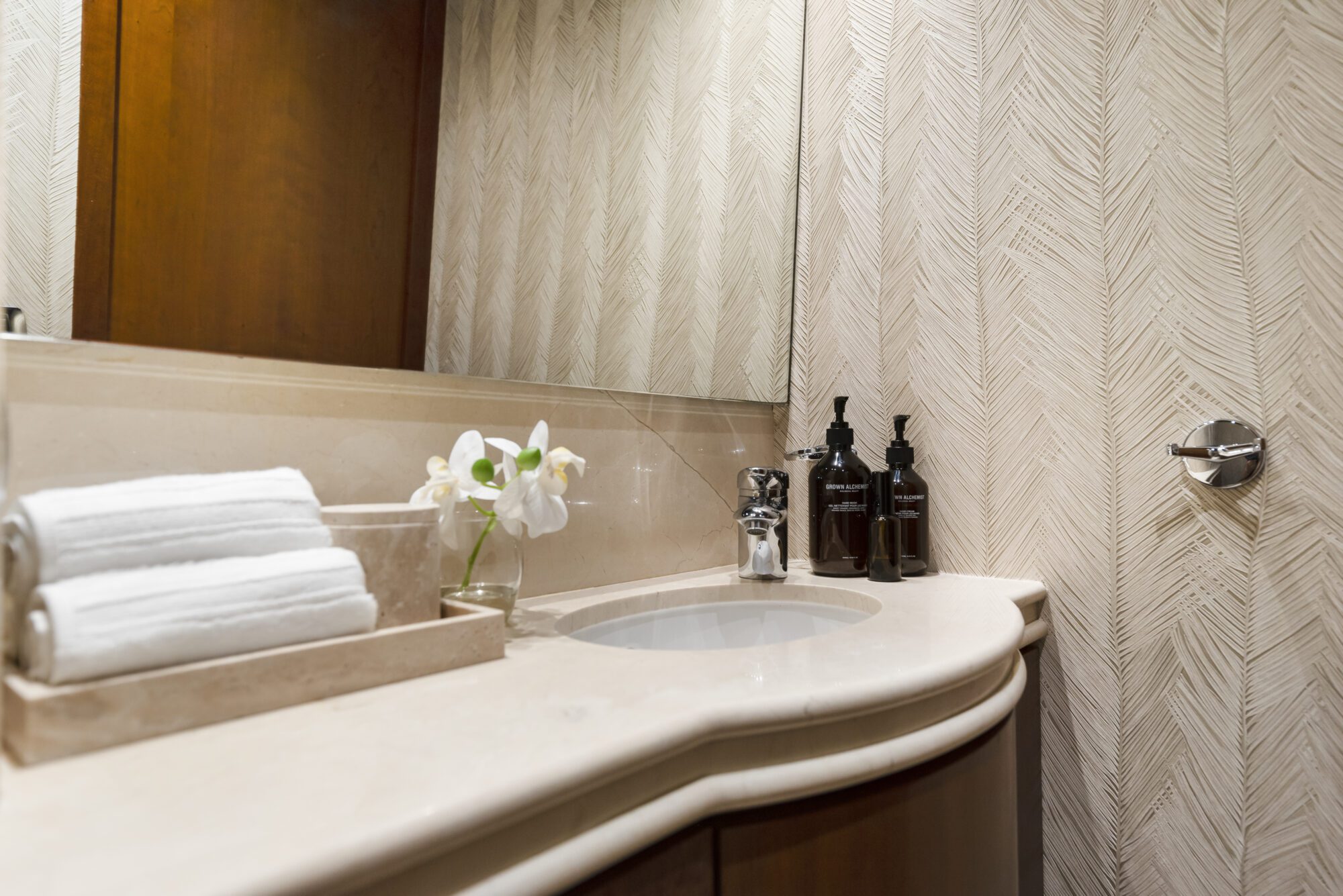 Modern bathroom countertop with a built-in sink, rolled white towels, a small vase with a white orchid, and dark soap dispensers. Light-colored patterned walls and a large mirror are visible.