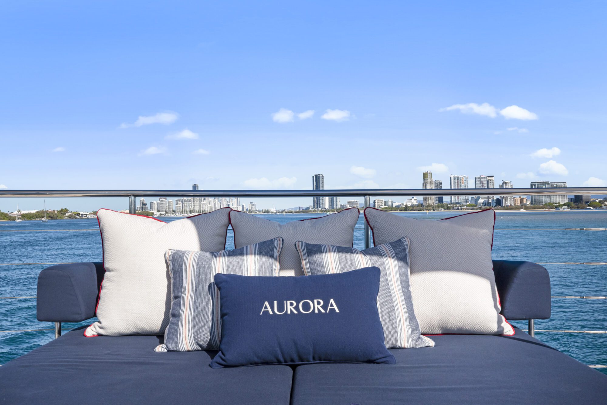 A cozy outdoor sofa with blue and white striped pillows sits on a yacht, with a pillow labeled "AURORA." In the background, a city skyline is visible across the blue water under a clear sky.
