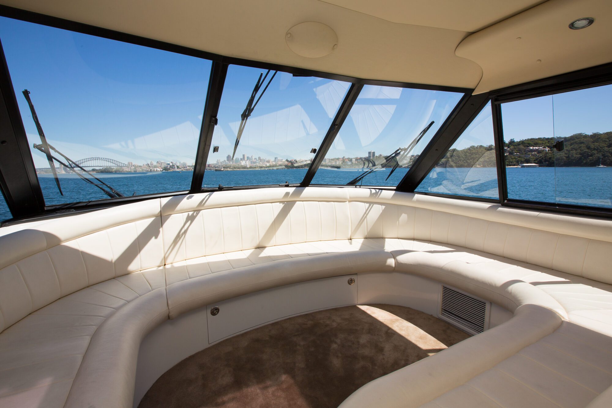 The image shows the interior of a yacht with white curved seating and large windows, providing a clear view of blue water and a distant city skyline under a sunny sky.
