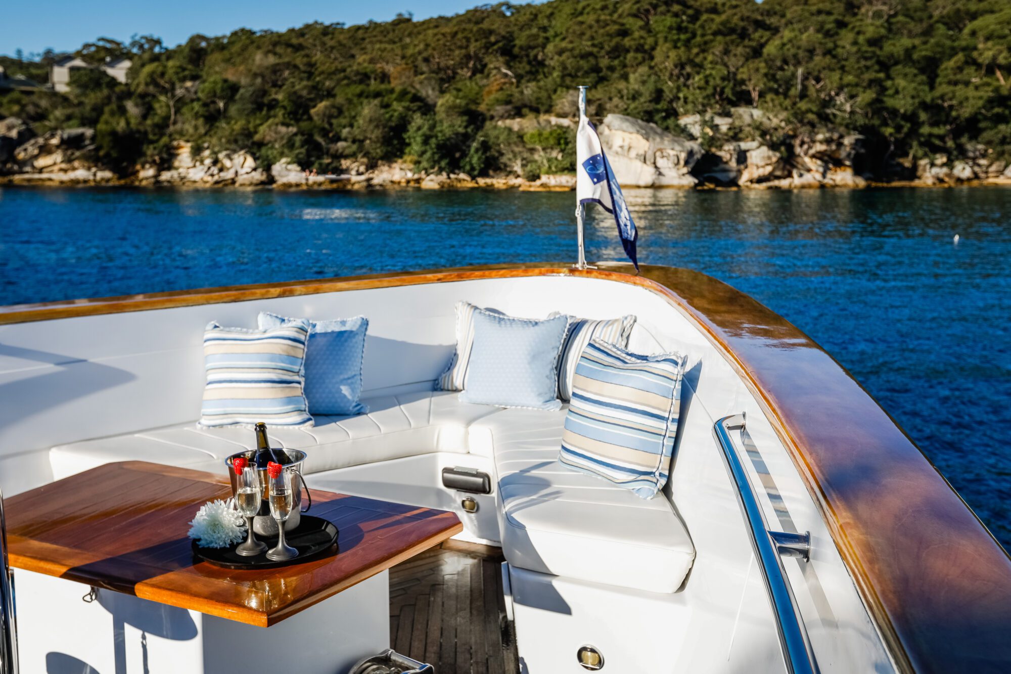 The bow of a luxury yacht features white cushioned seating with blue and striped pillows, a small table with champagne and glasses, overlooking calm blue water and a tree-lined shore in the background.